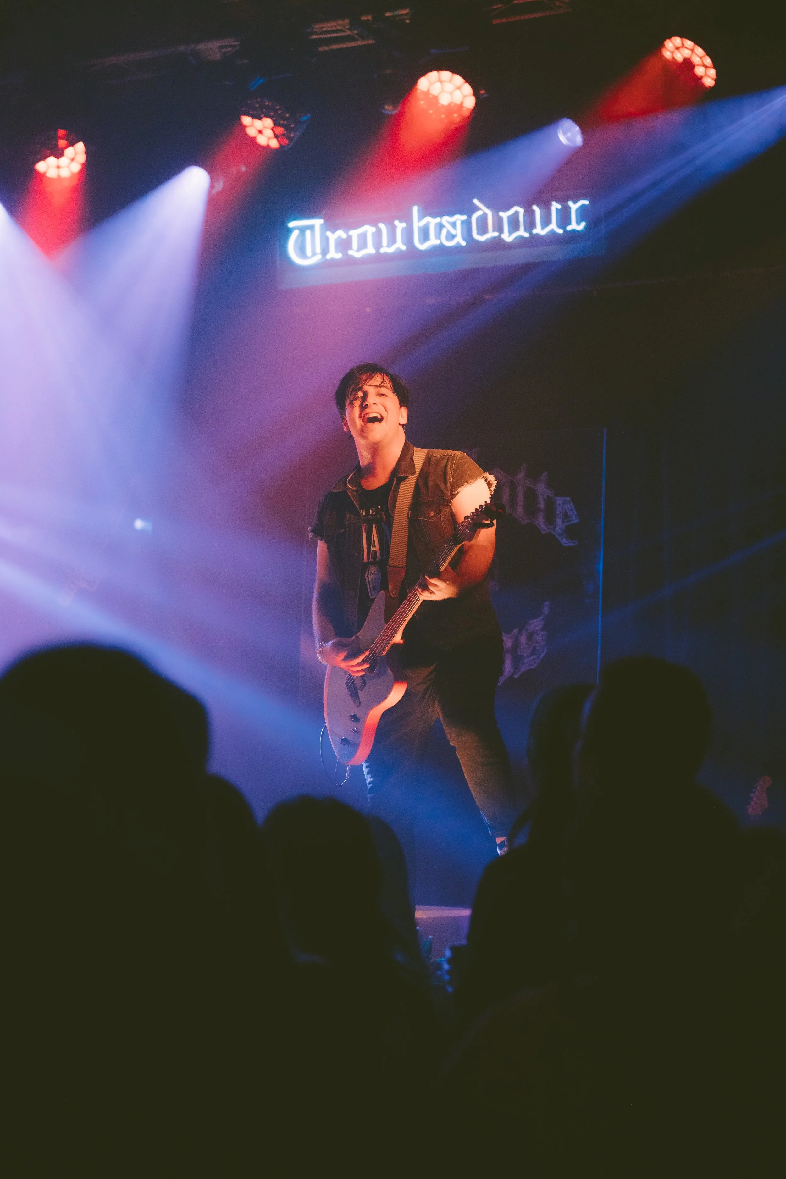 A musician is performing on stage with a guitar, laughing or singing, under colorful stage lighting with a "Troubadour" sign above him. Audience members are visible in the foreground.