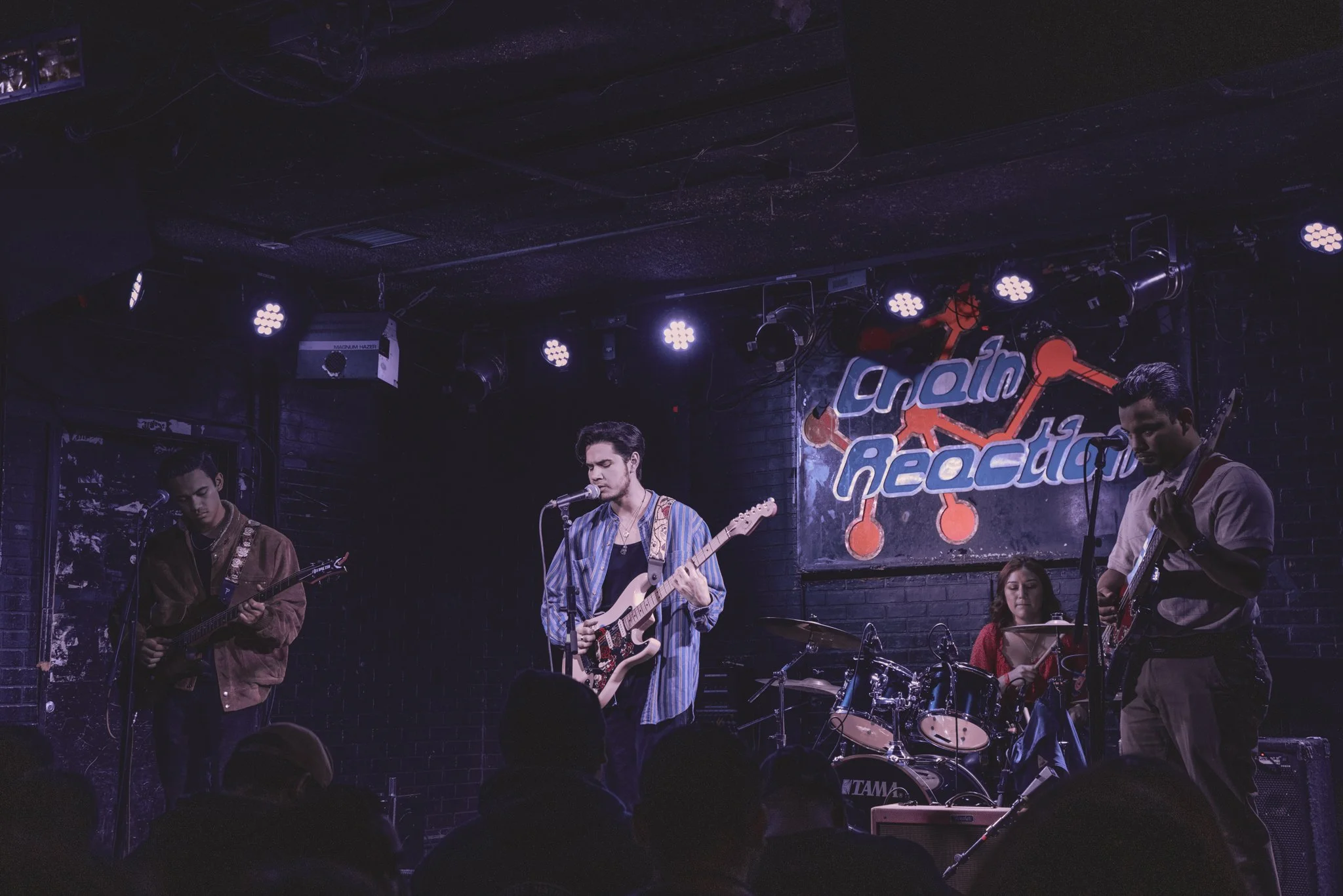 A band performing live on stage at a venue called 'Clash Addiction,' with four members playing guitars and drums under purple stage lights.