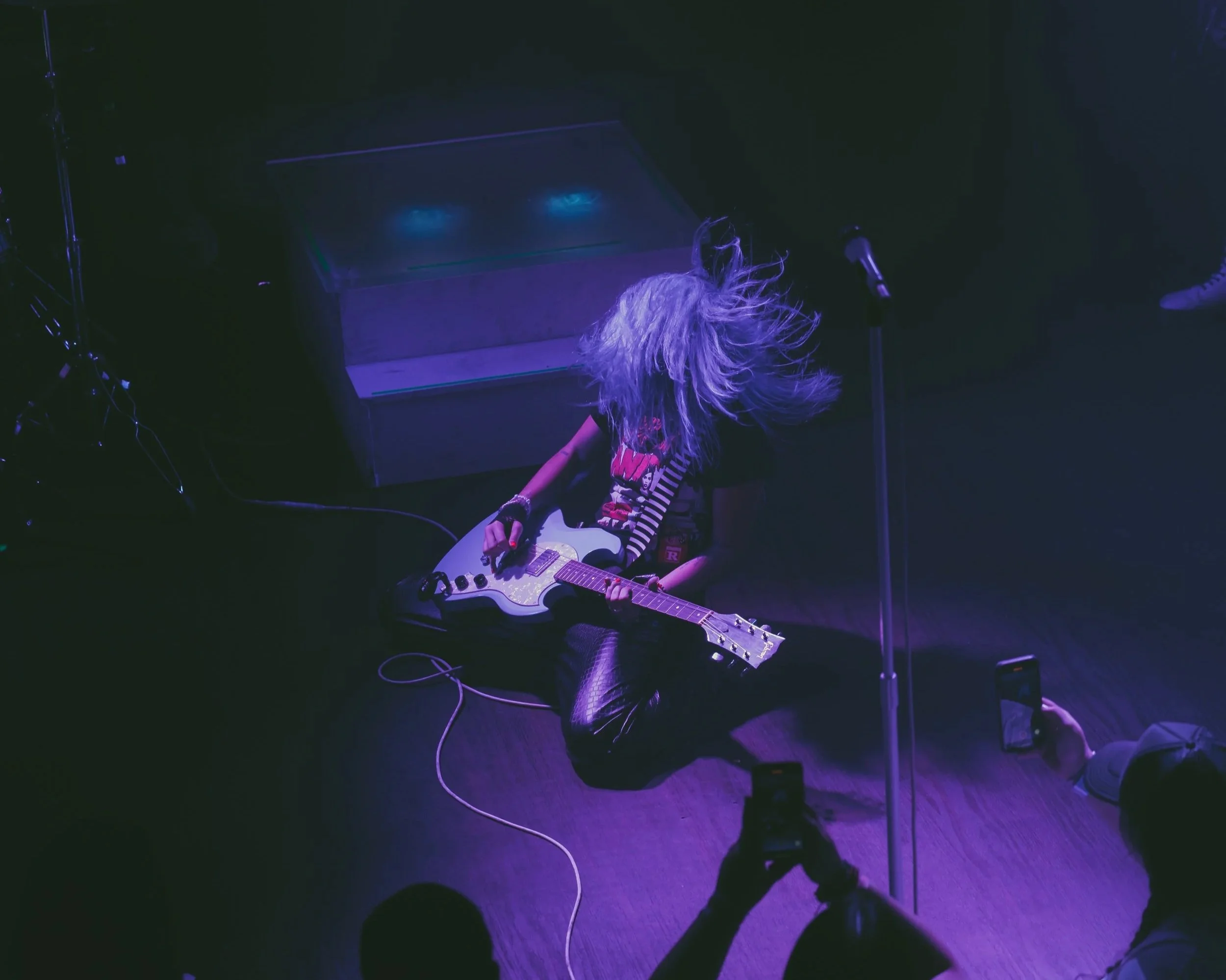 Female musician with long, flowing hair playing an electric guitar on stage, illuminated by purple lighting, with audience members taking photos.