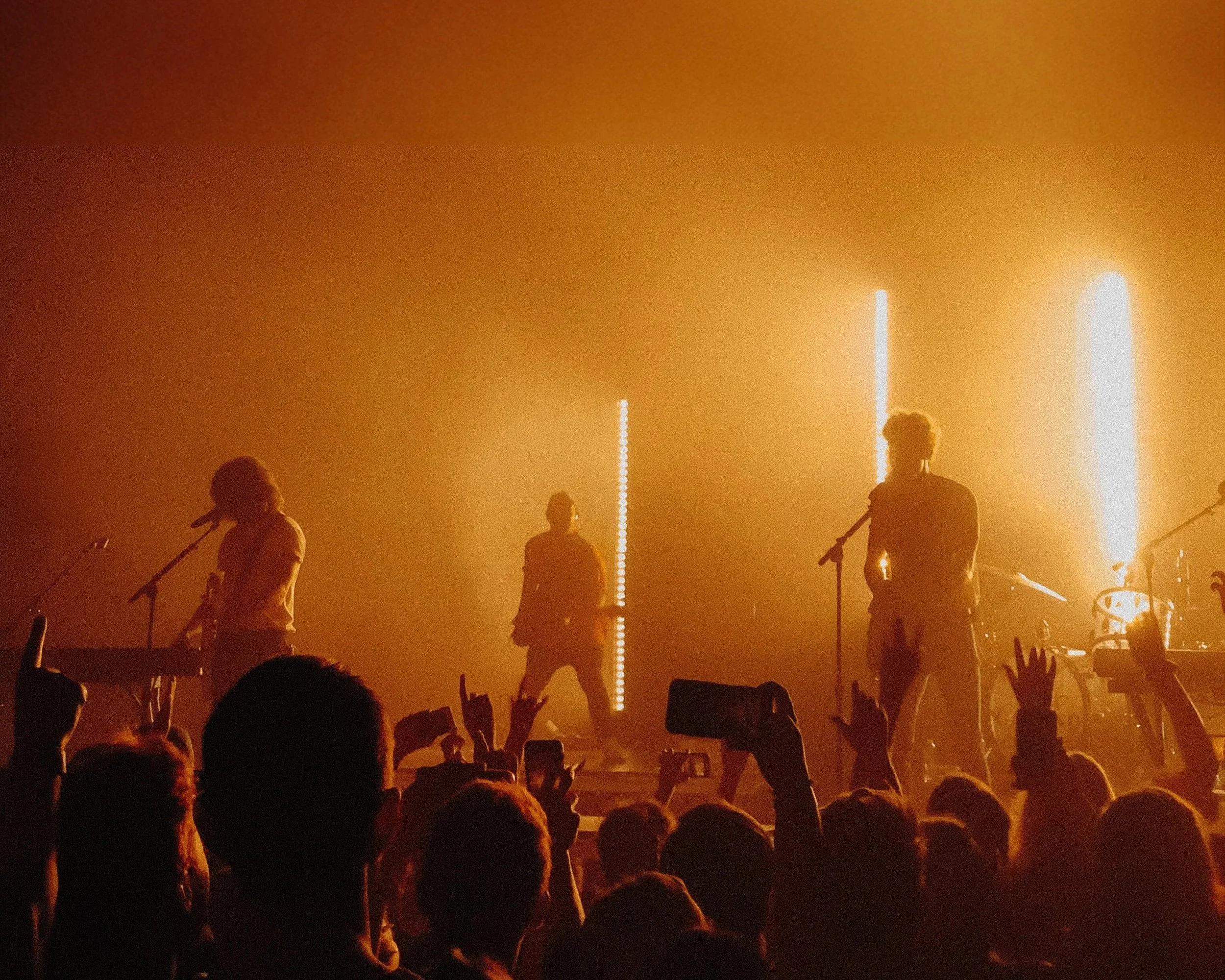 A band performing on stage with orange lighting, featuring silhouettes of musicians and audience members with hands raised and some holding up phones.