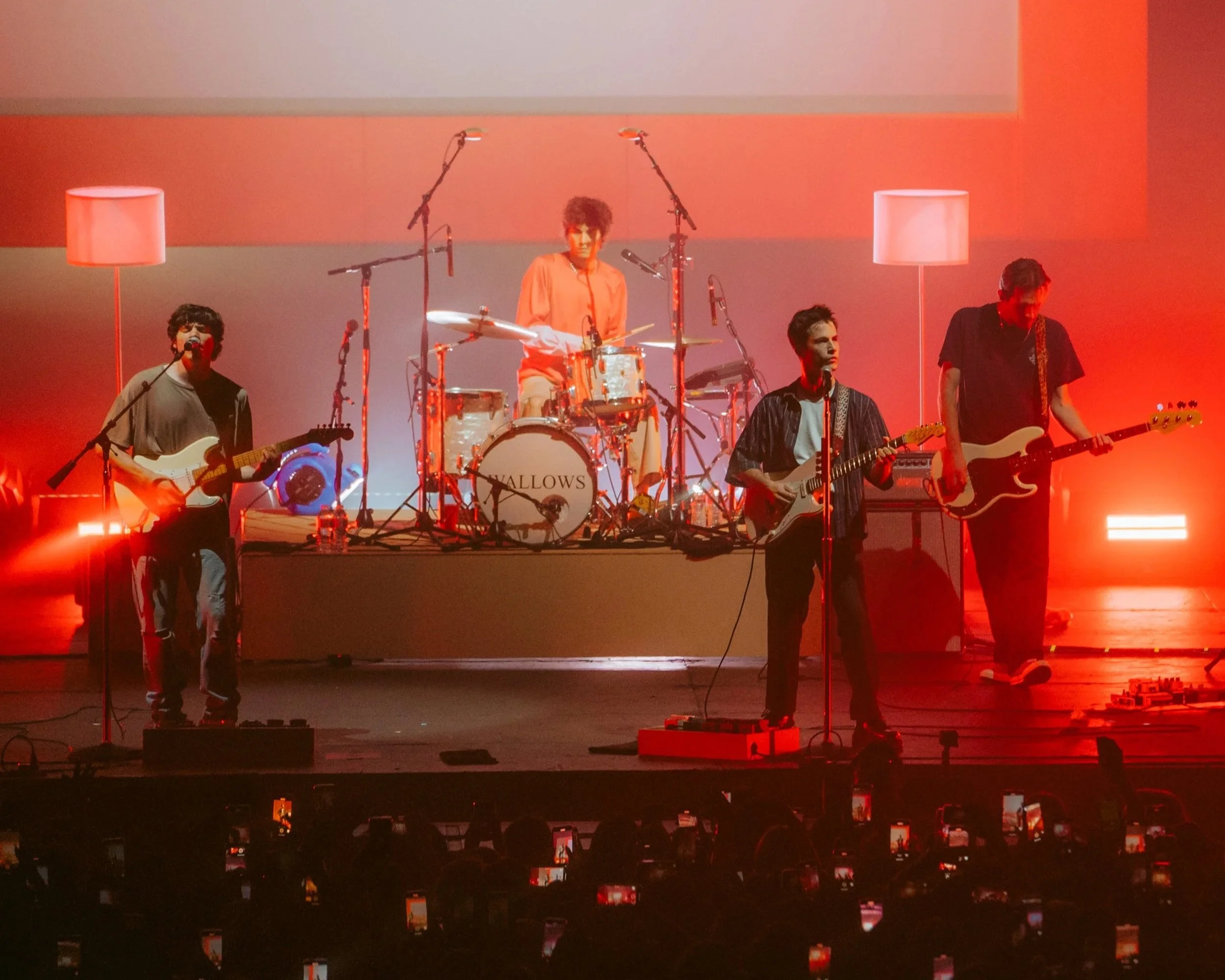 A band performing on stage with four members playing guitars and drums, with a background of red and orange lighting and two floor lamps.