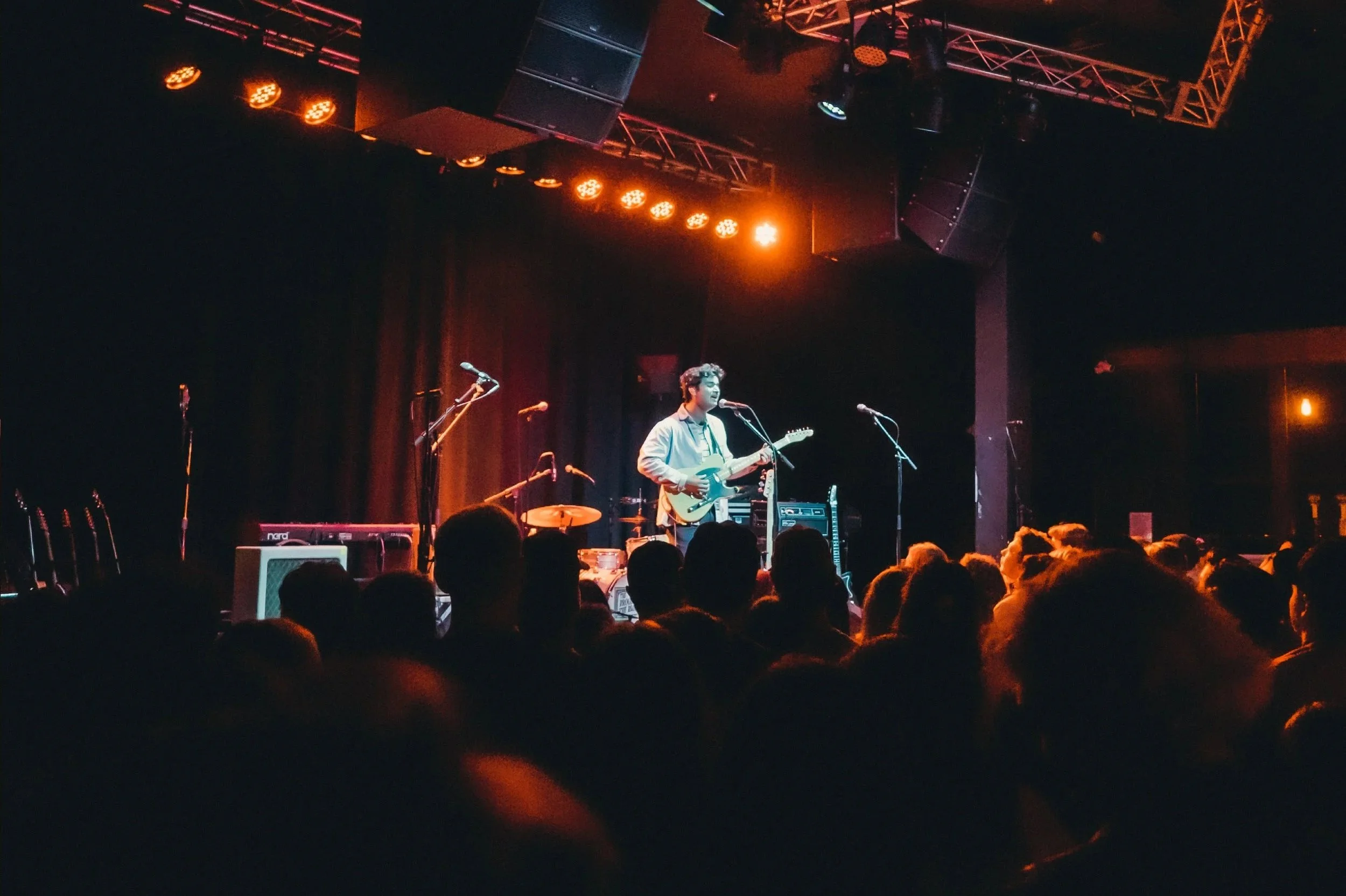 A musician performing on stage with an electric guitar, surrounded by microphones, with a dark banner background and stage lighting, and an audience watching.