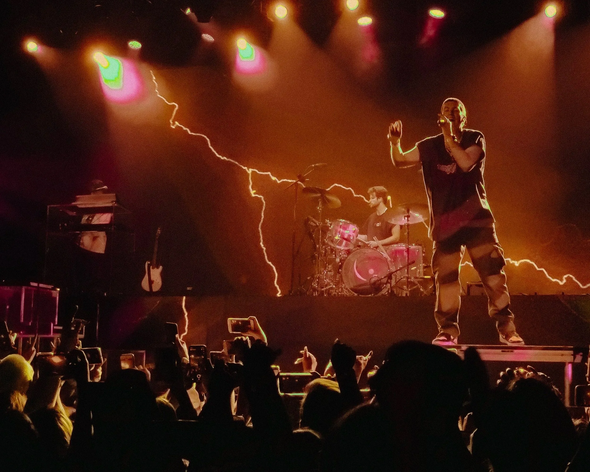 A concert scene with a singer at the microphone, a drummer in the background, and colorful lightning effects on the backdrop, with an audience taking photos and videos.