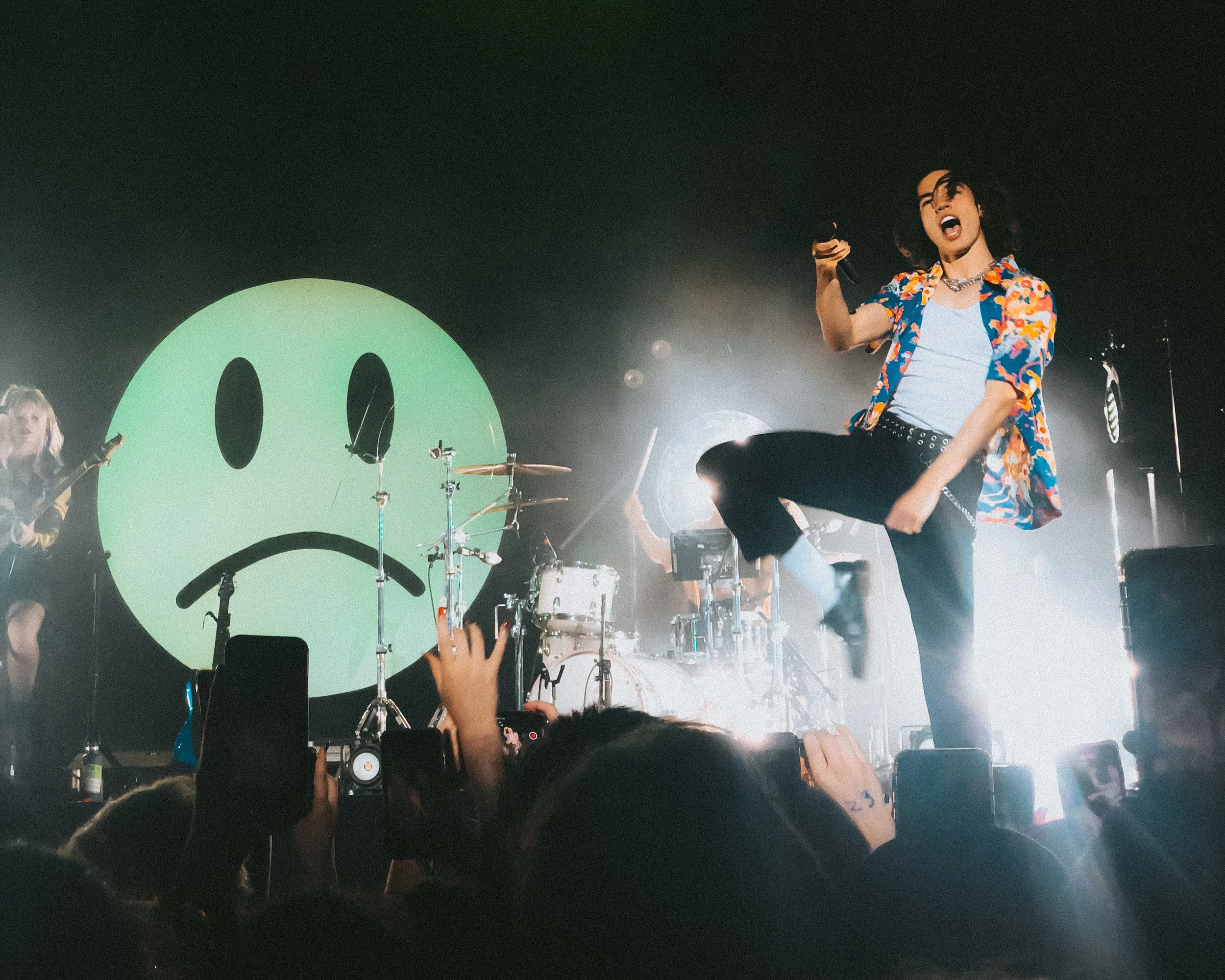 A performer singing passionately on stage, wearing a colorful floral shirt, black pants, and a choker, with a band behind them, a giant smiley face on the backdrop, and an enthusiastic crowd taking photos and videos.