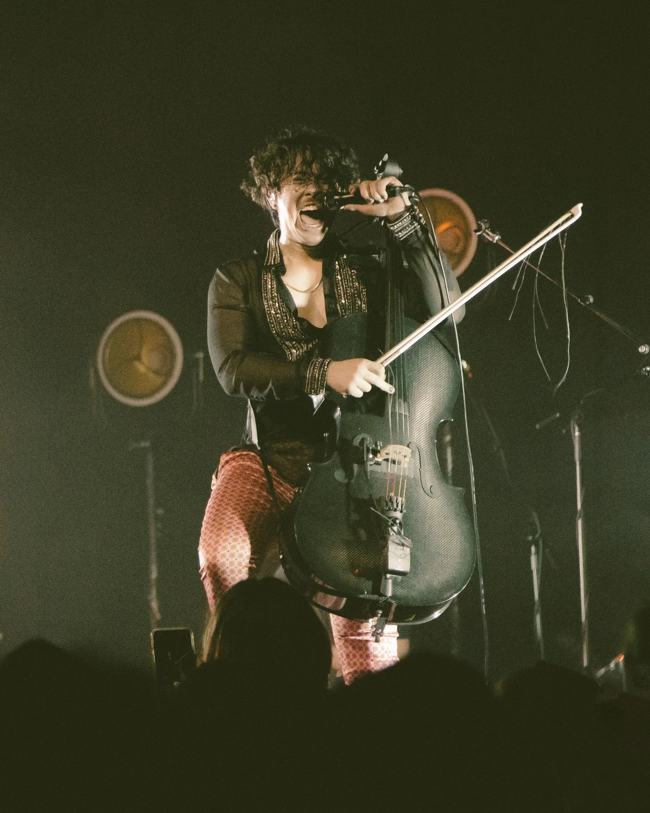 A musician passionately performing on stage with a microphone and a cello, wearing a black jacket and reddish pants, surrounded by stage lights.