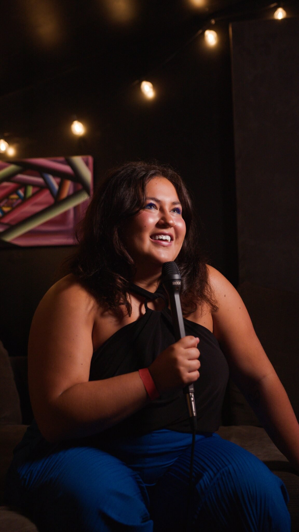 A woman with curly hair, wearing a black top and blue pants, smiling and holding a microphone while seated in a dimly lit space with string lights overhead.