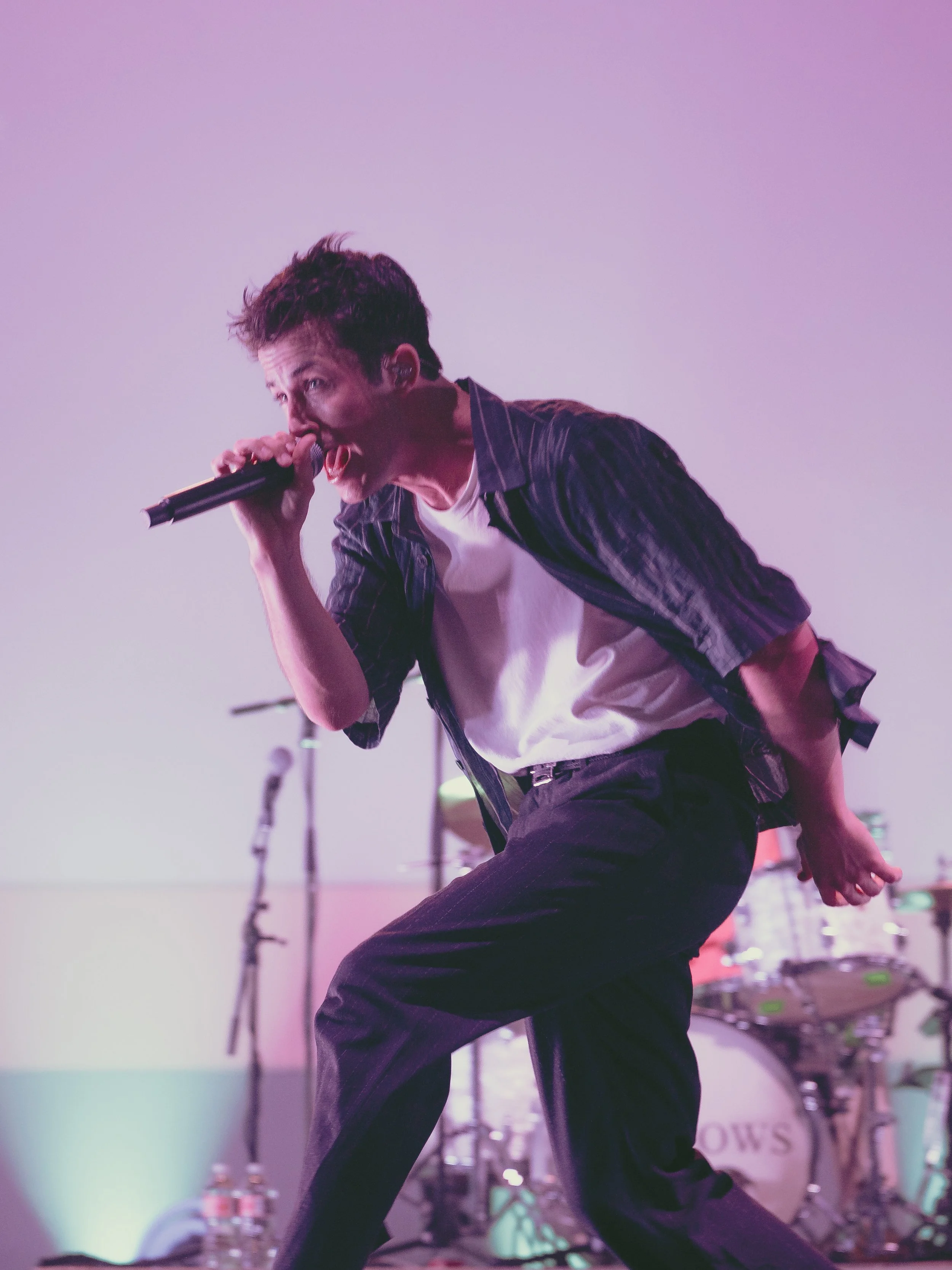 A male singer passionately performs on stage holding a microphone, wearing a white t-shirt and a dark striped shirt. The background shows musical instruments and stage equipment.