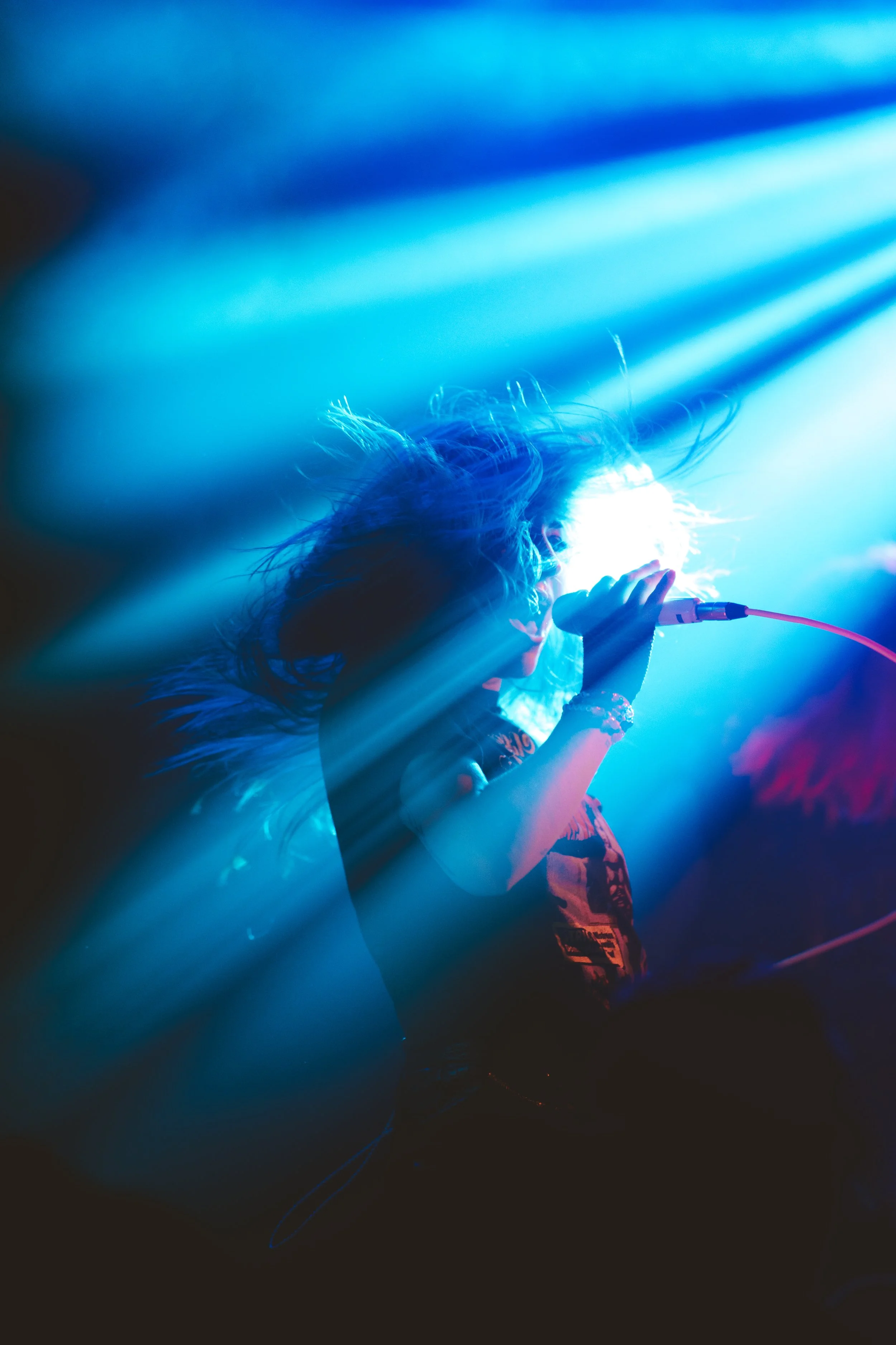 A singer performing on stage with blue and purple stage lighting, holding a microphone, with hair flying in the air.