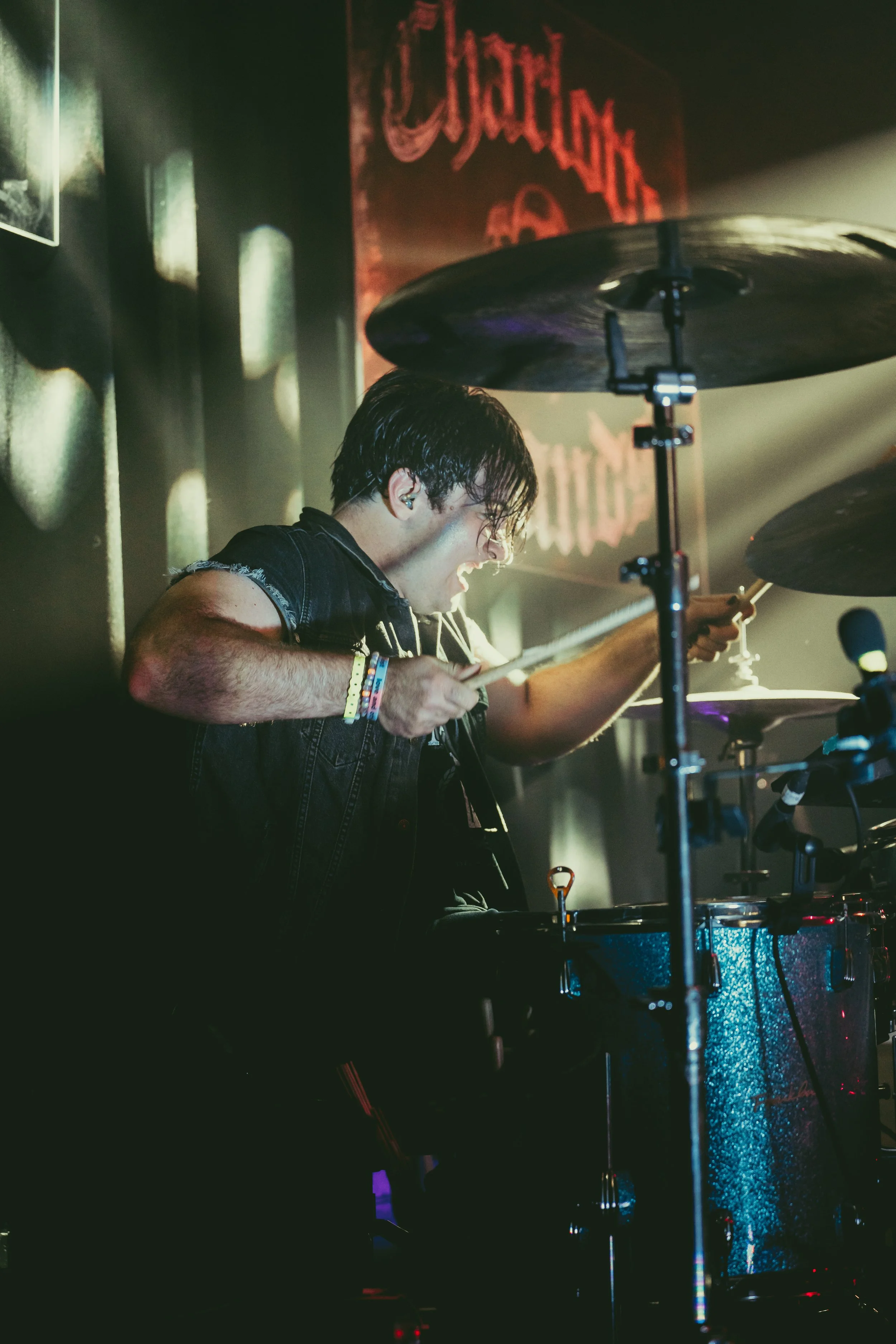 A young man playing drums passionately during a live music performance, with colorful stage lighting and a dark background featuring a red sign.