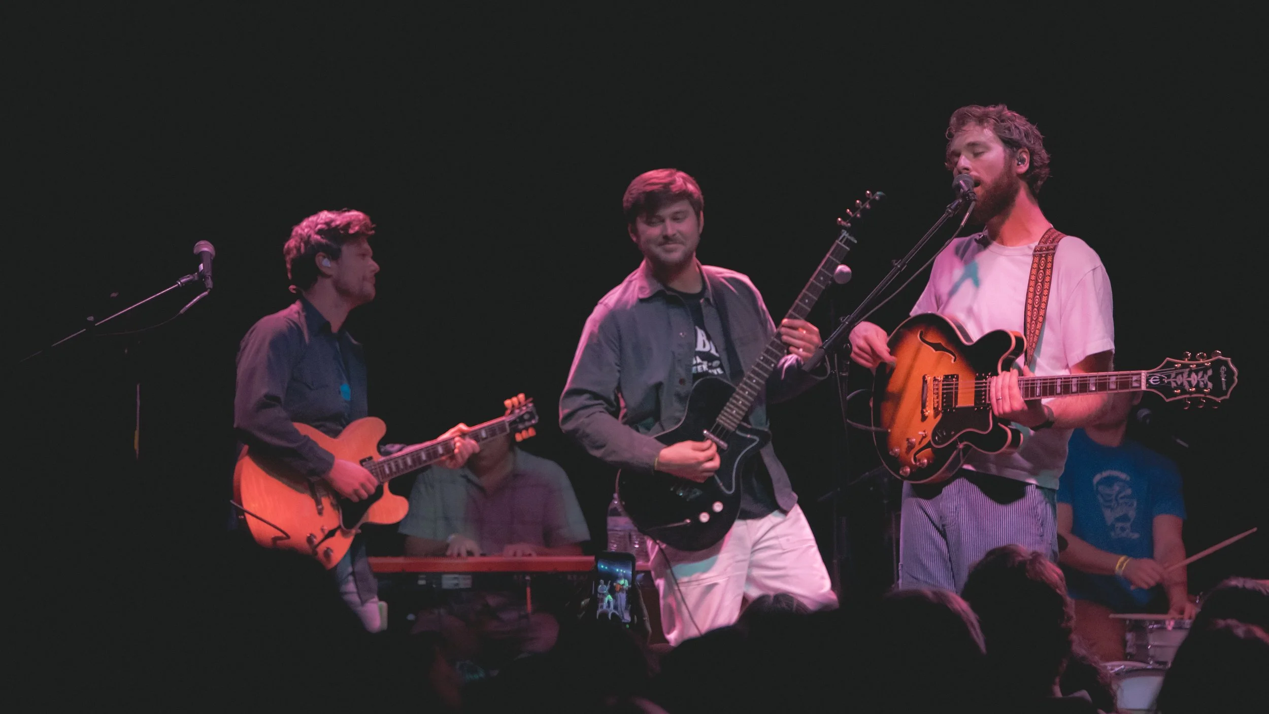 Band performing on stage with three guitarists, one keyboard player, and a drummer, under stage lighting.