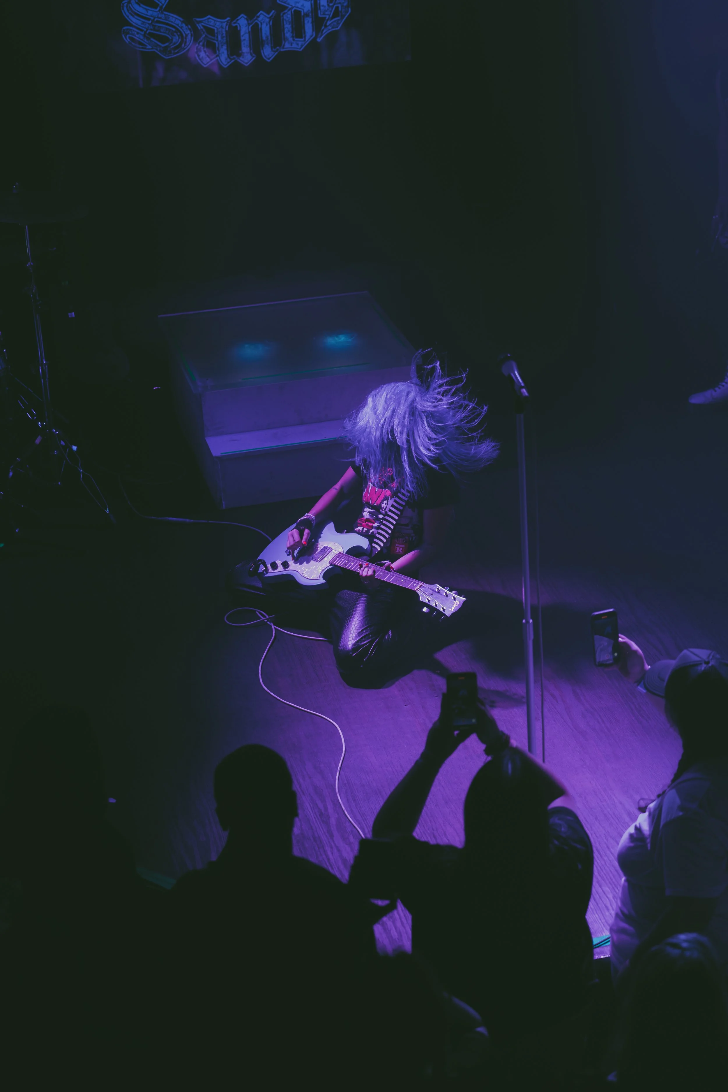 A female guitarist with long, wavy hair, sitting cross-legged on stage, playing an electric guitar during a live performance, illuminated by purple and blue stage lights, with audience members taking photos.