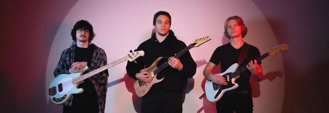 Three young men standing with guitars, posing against a plain background with lighting creating a shadow behind them.