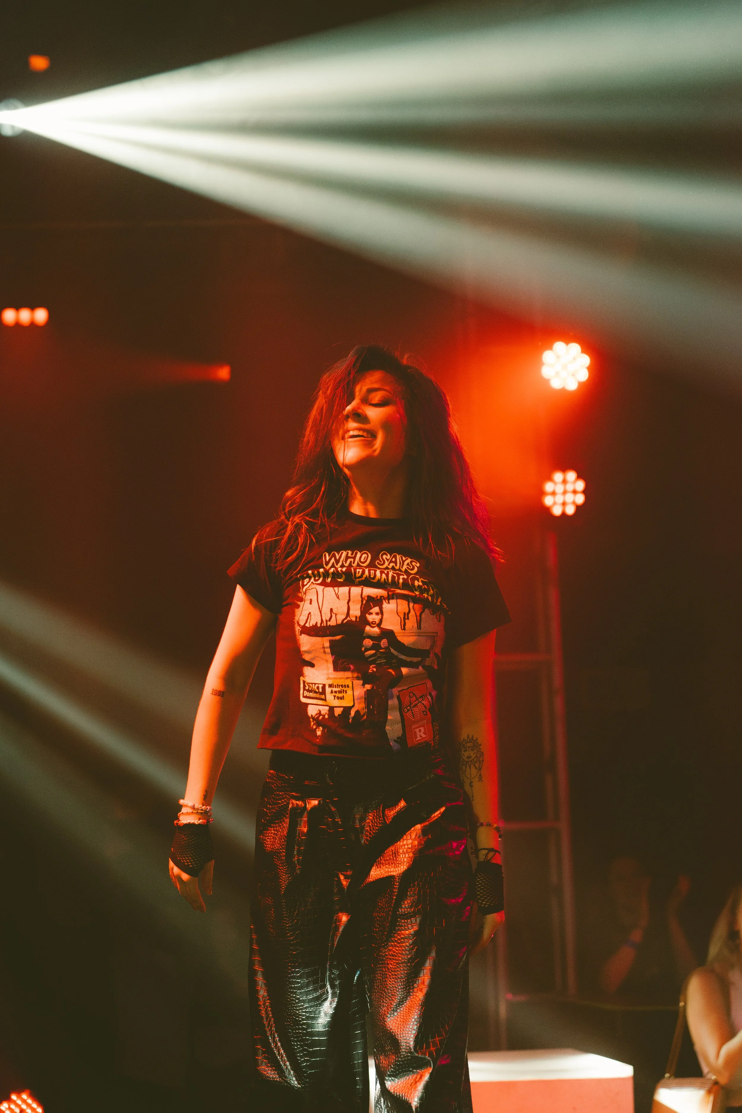 A woman with long, dark, wavy hair smiling and dancing on a stage illuminated by red and white lights, wearing a graphic black T-shirt and shiny pants.