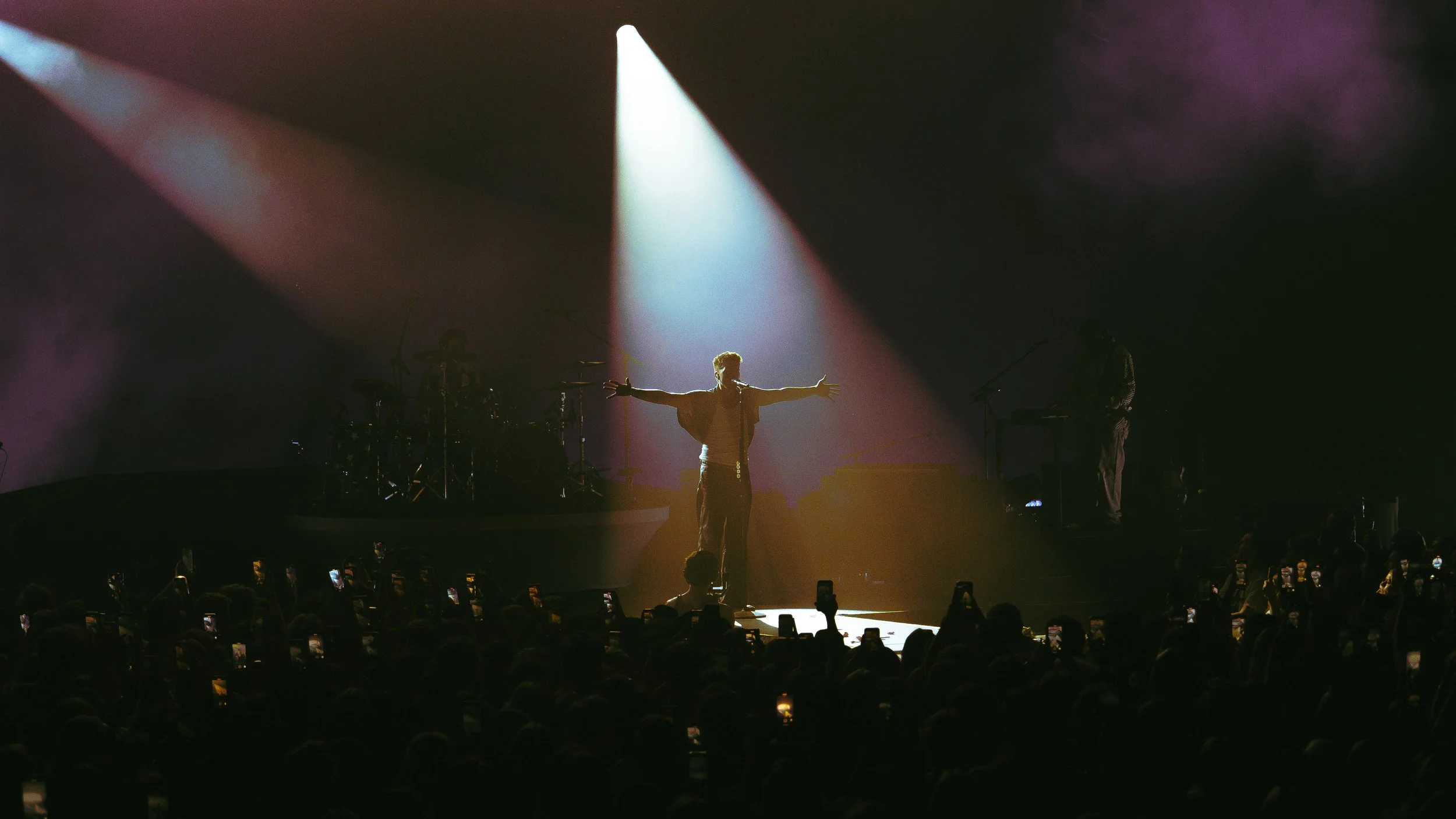 A performer standing on stage with arms outstretched, illuminated by a spotlight, during a concert with an audience taking photos.