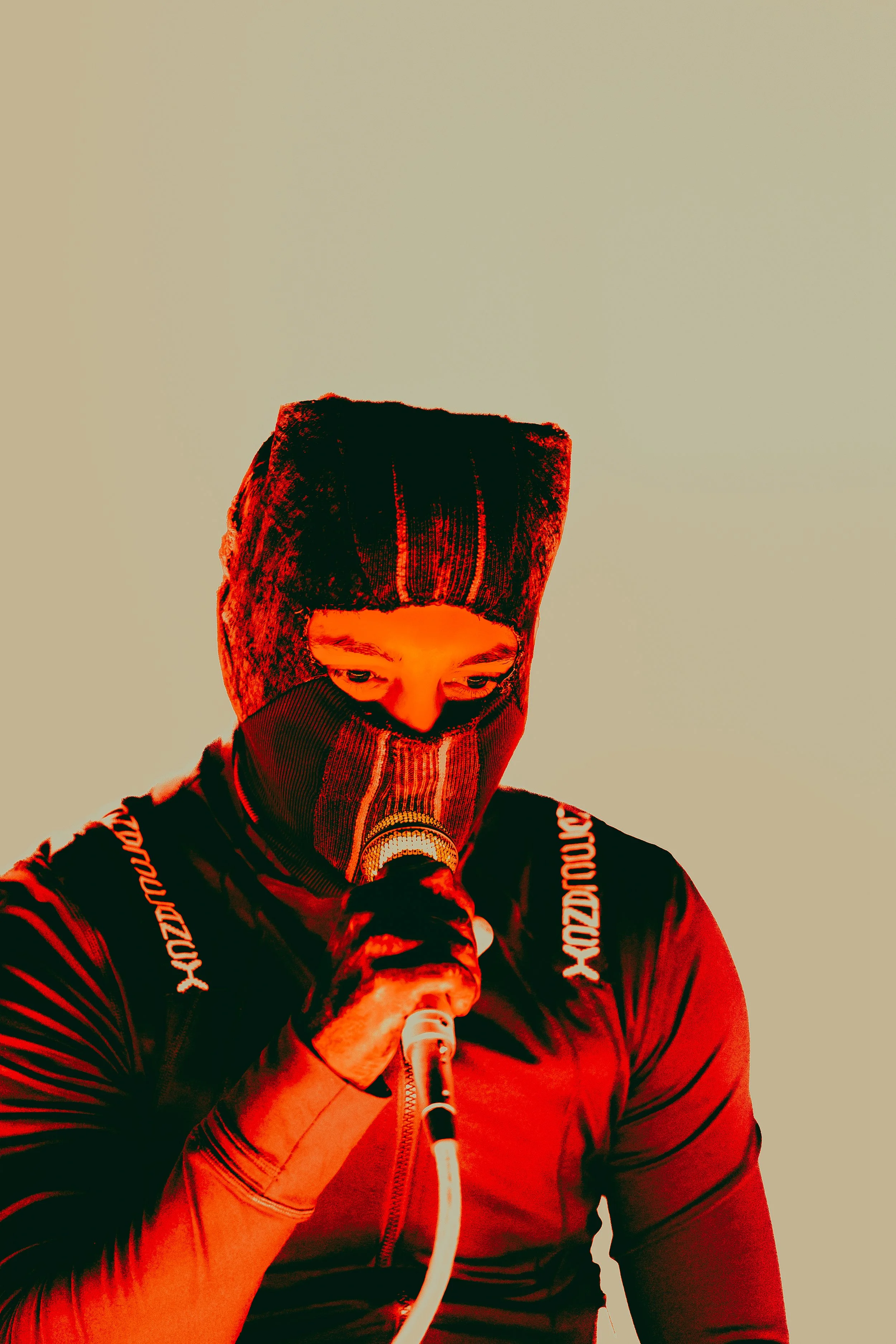 Person wearing a ski mask or face covering, holding a microphone, and dressed in a black jacket with red lighting, against a plain background.