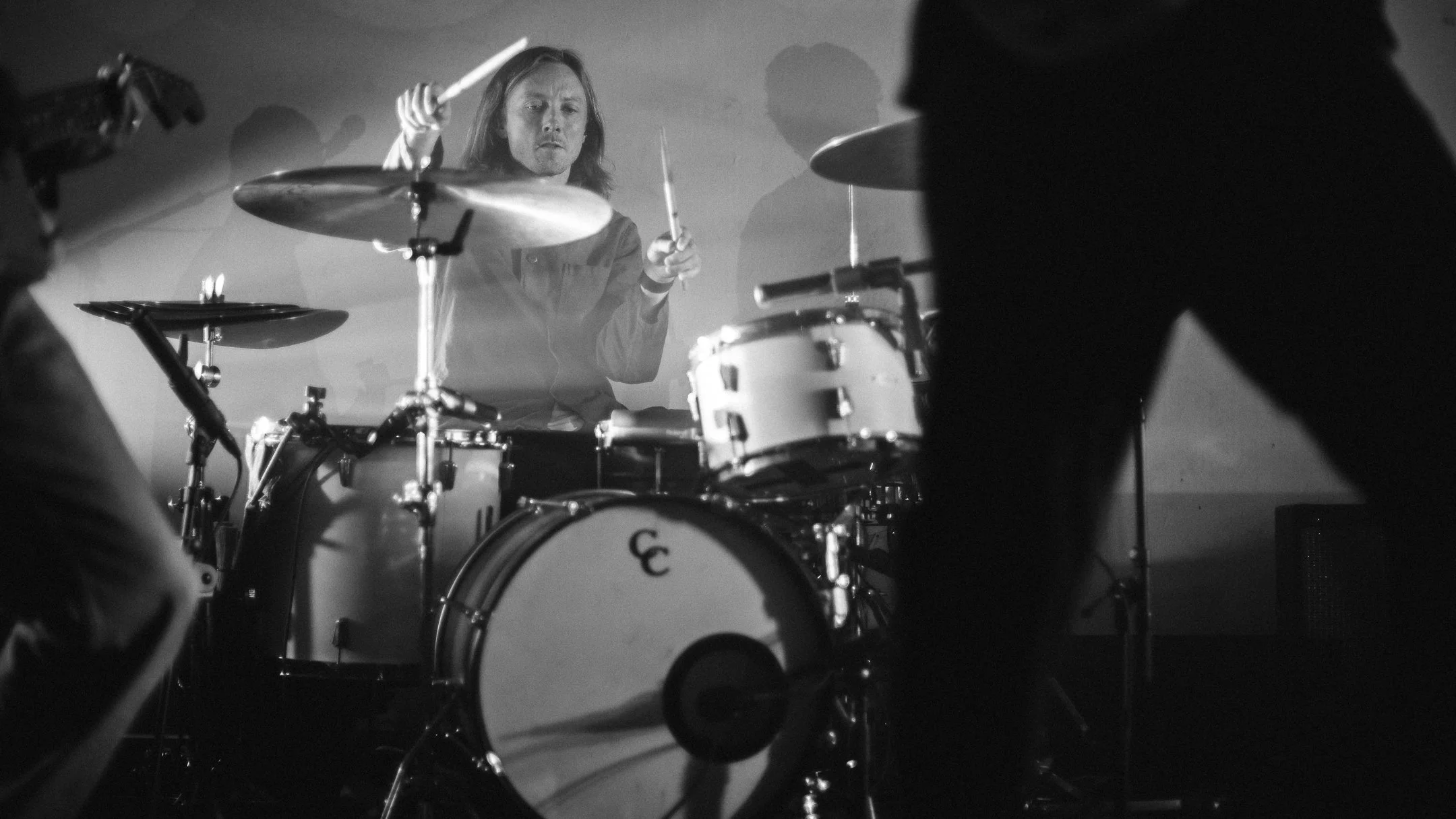 A black and white photo of a man playing drums on stage, with other musicians partially visible in the foreground and background.