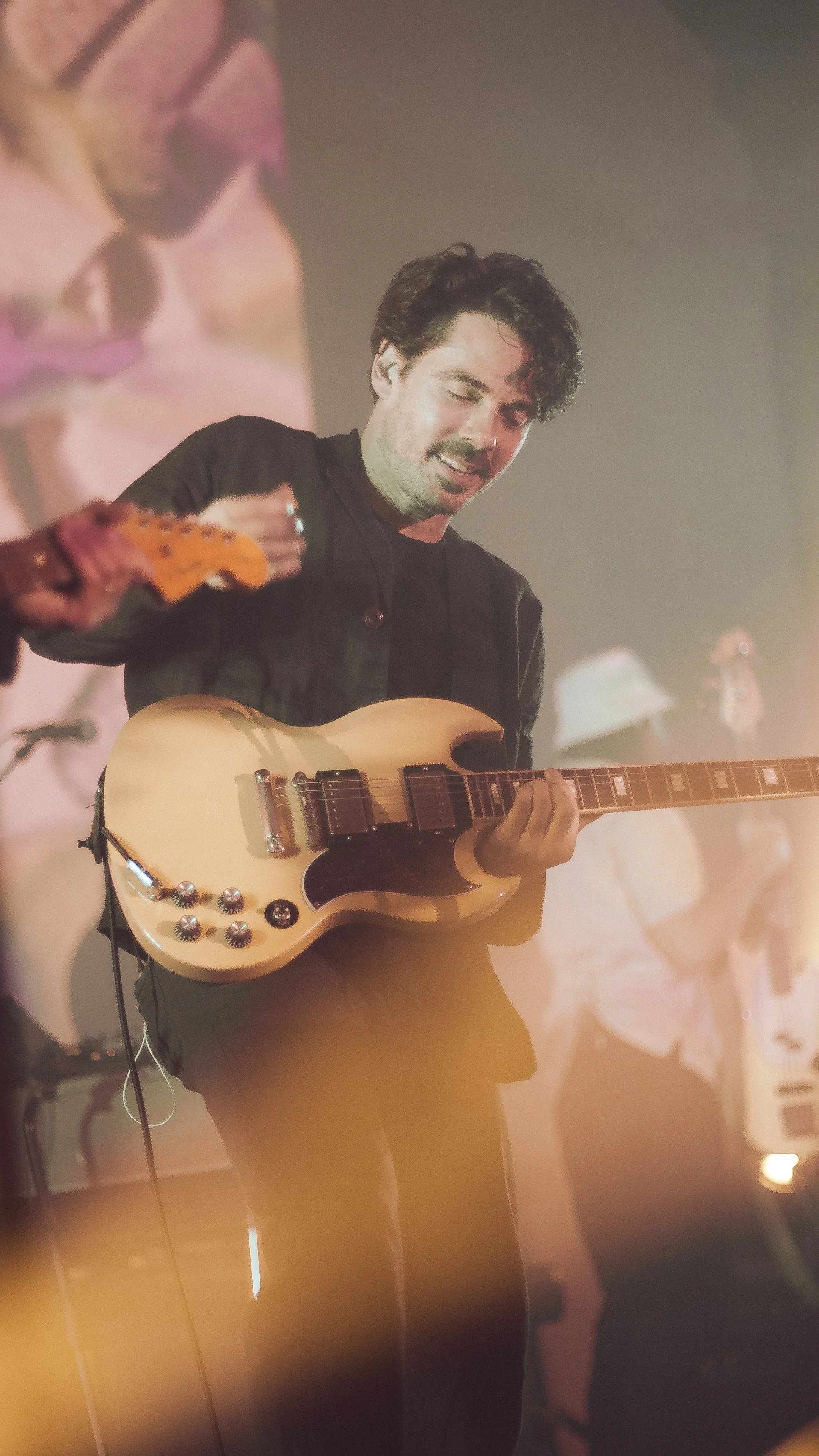 A man playing a cream-colored electric guitar on stage with a dark background and colorful lights.