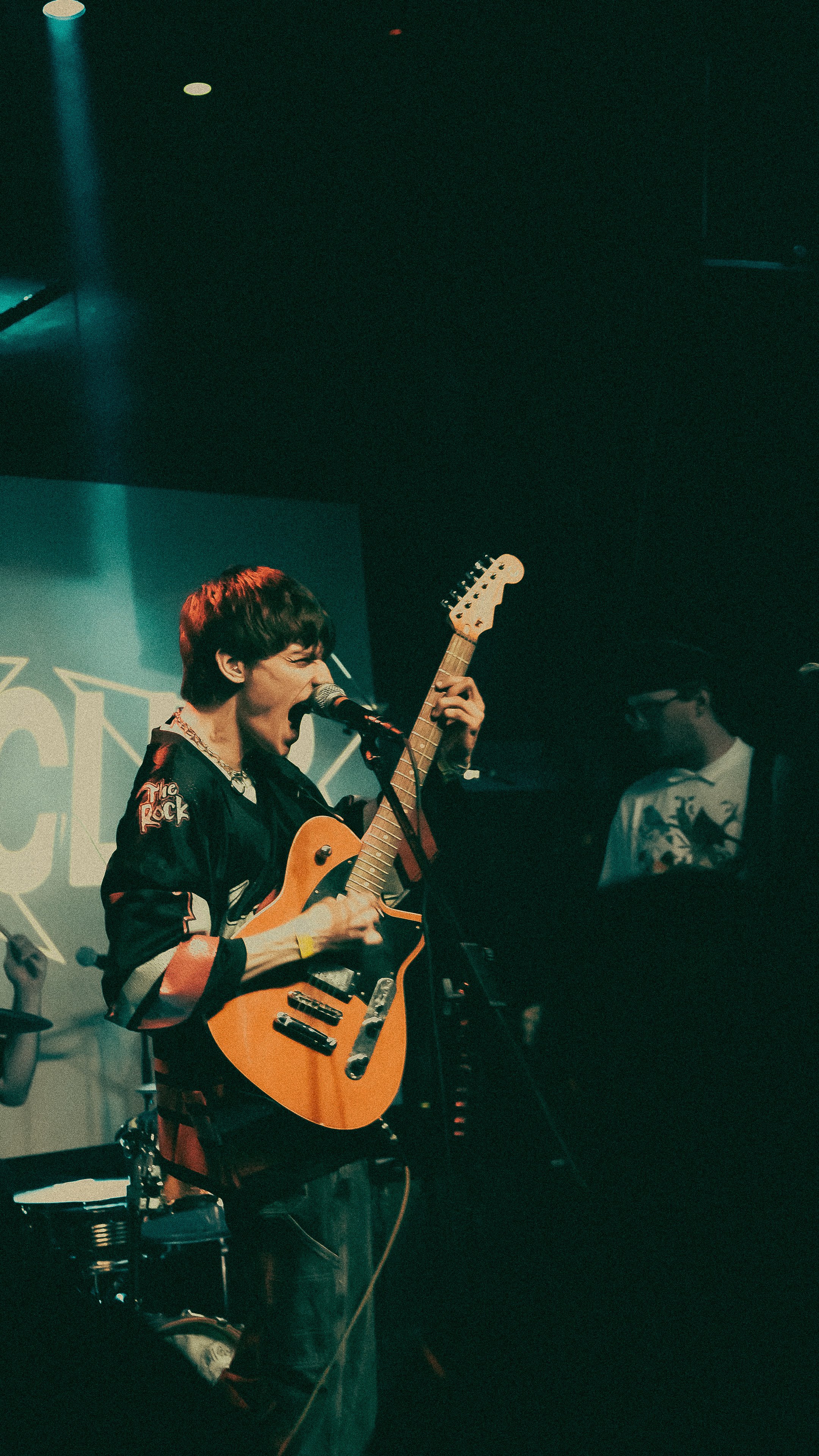 A young male musician singing into a microphone while playing an electric guitar on stage, with a person in the background.