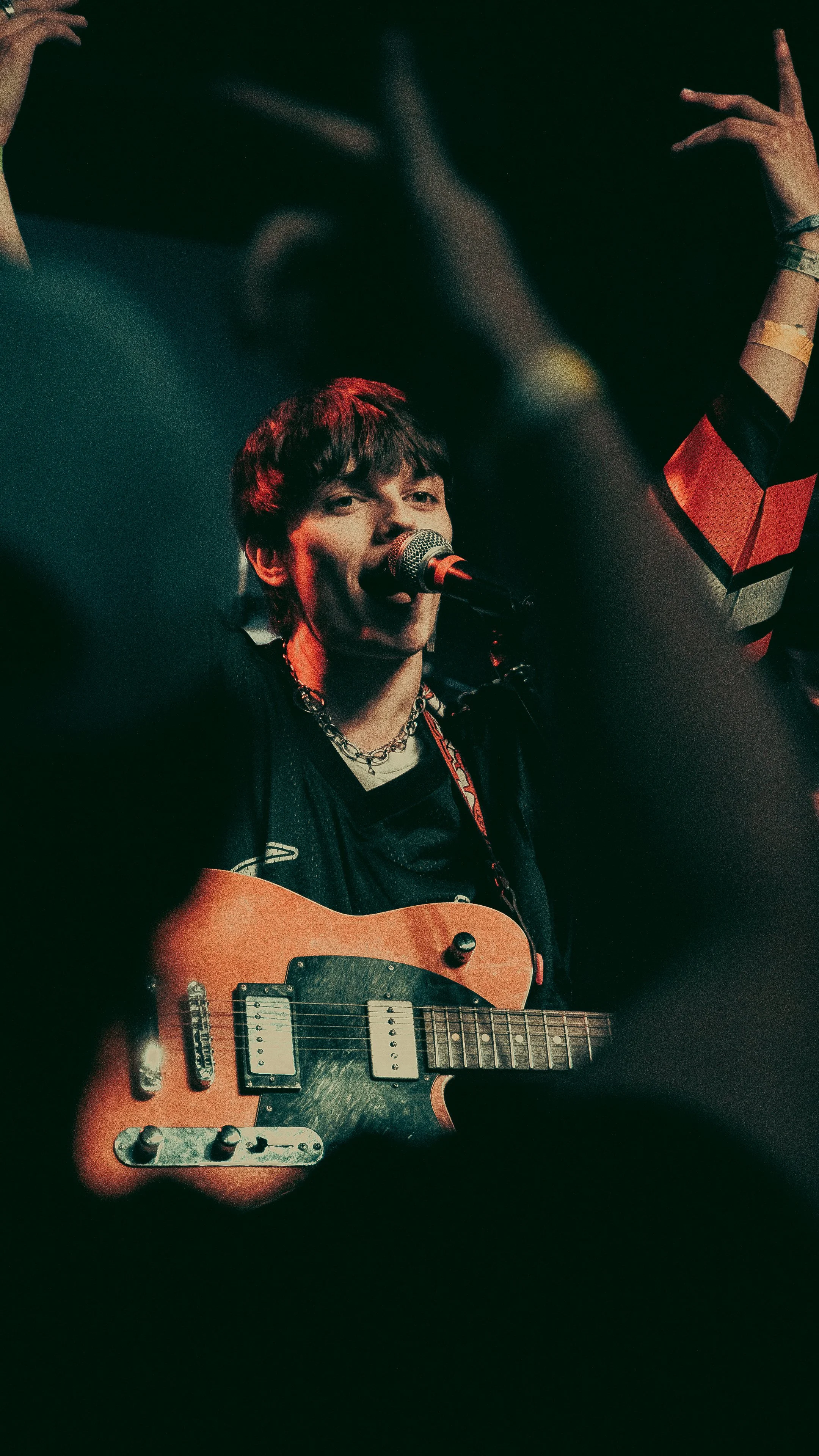 A young male musician singing into a microphone while playing an electric guitar on stage, surrounded by dark lighting.