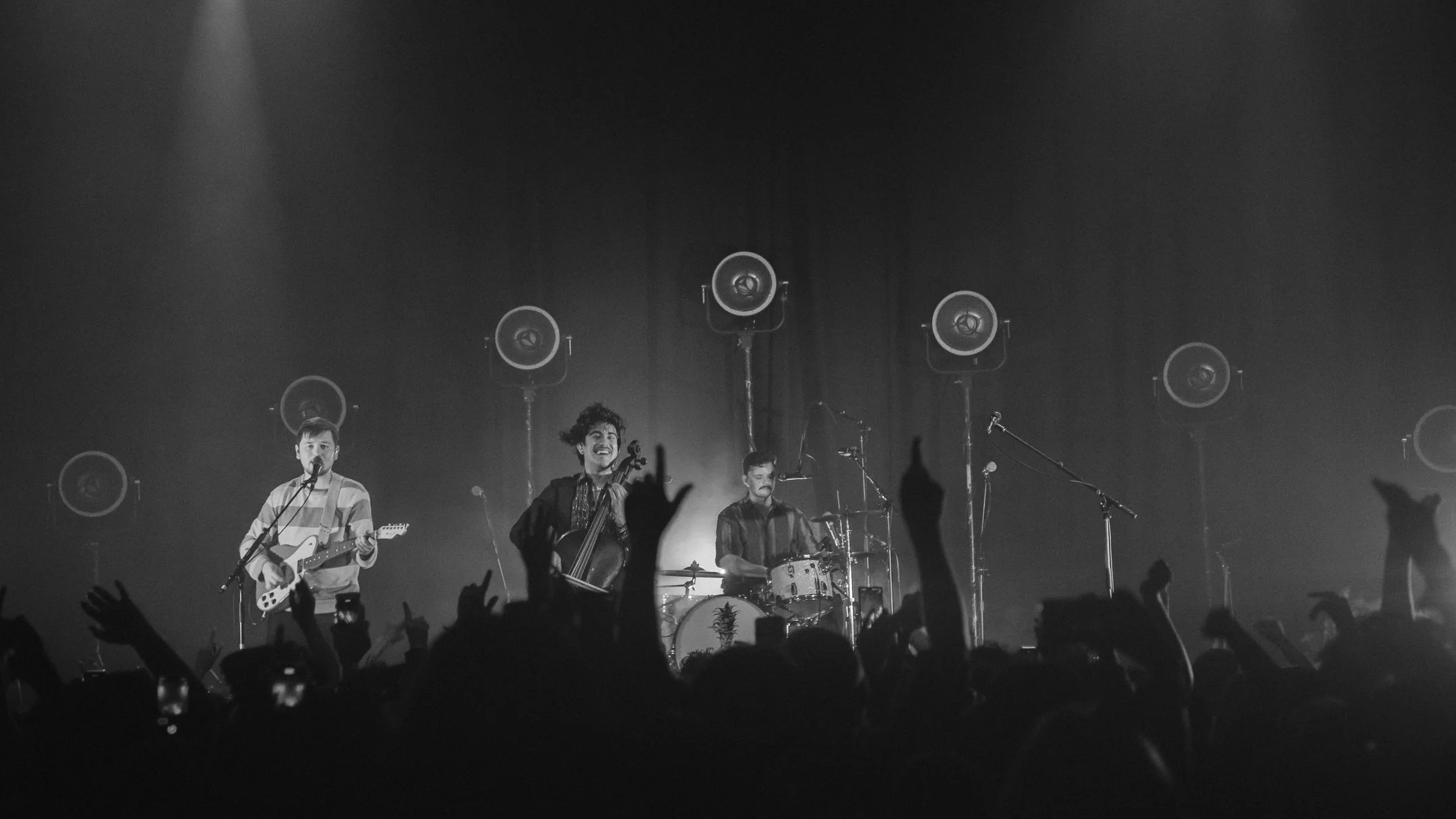 Black and white photo of a band performing on stage, with 3 band members playing instruments and singing, audience with raised hands in front, stage lights shining down.