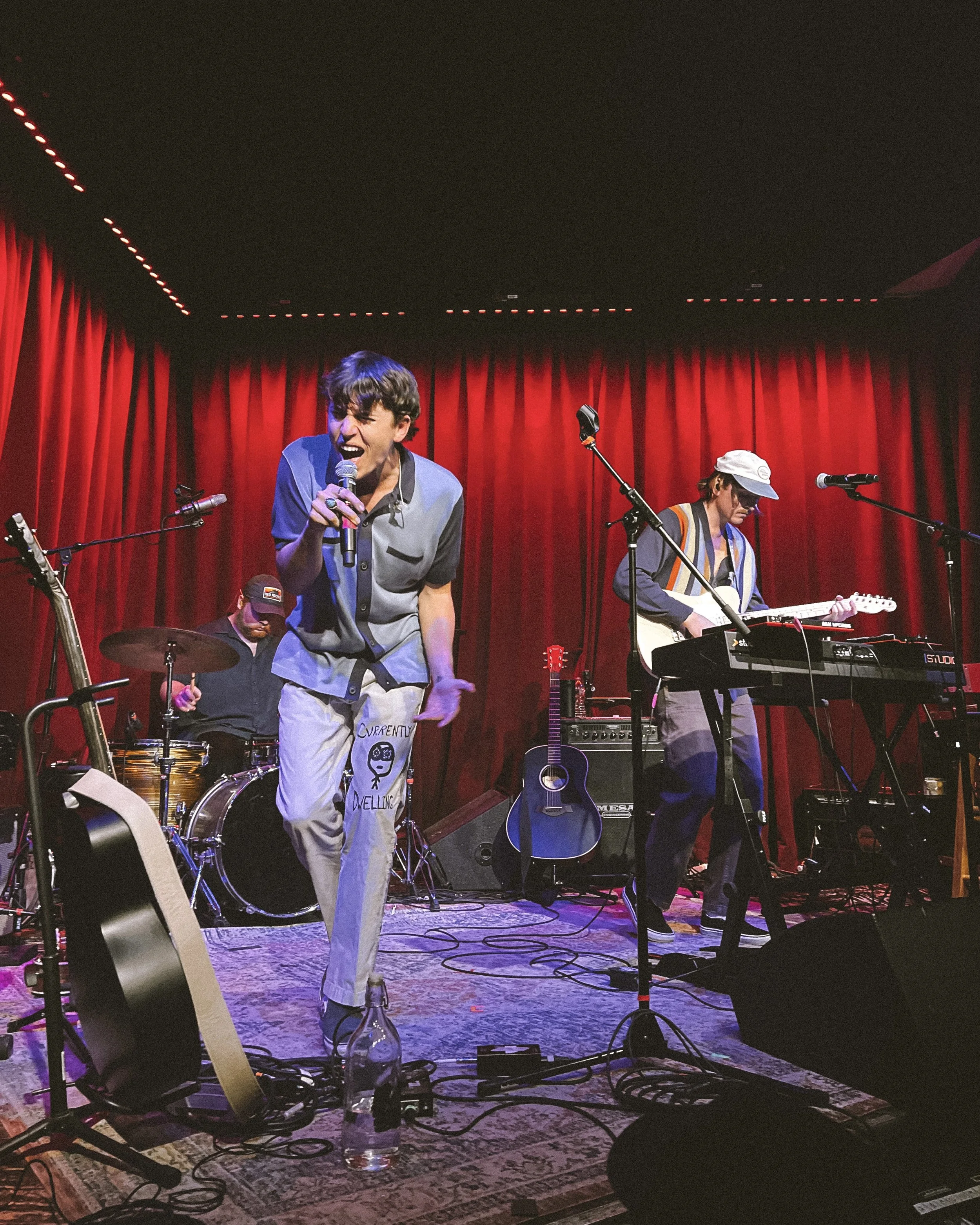 A band performing on stage with red curtains in the background, including a singer holding a microphone, a drummer, and a guitarist playing an electric guitar.