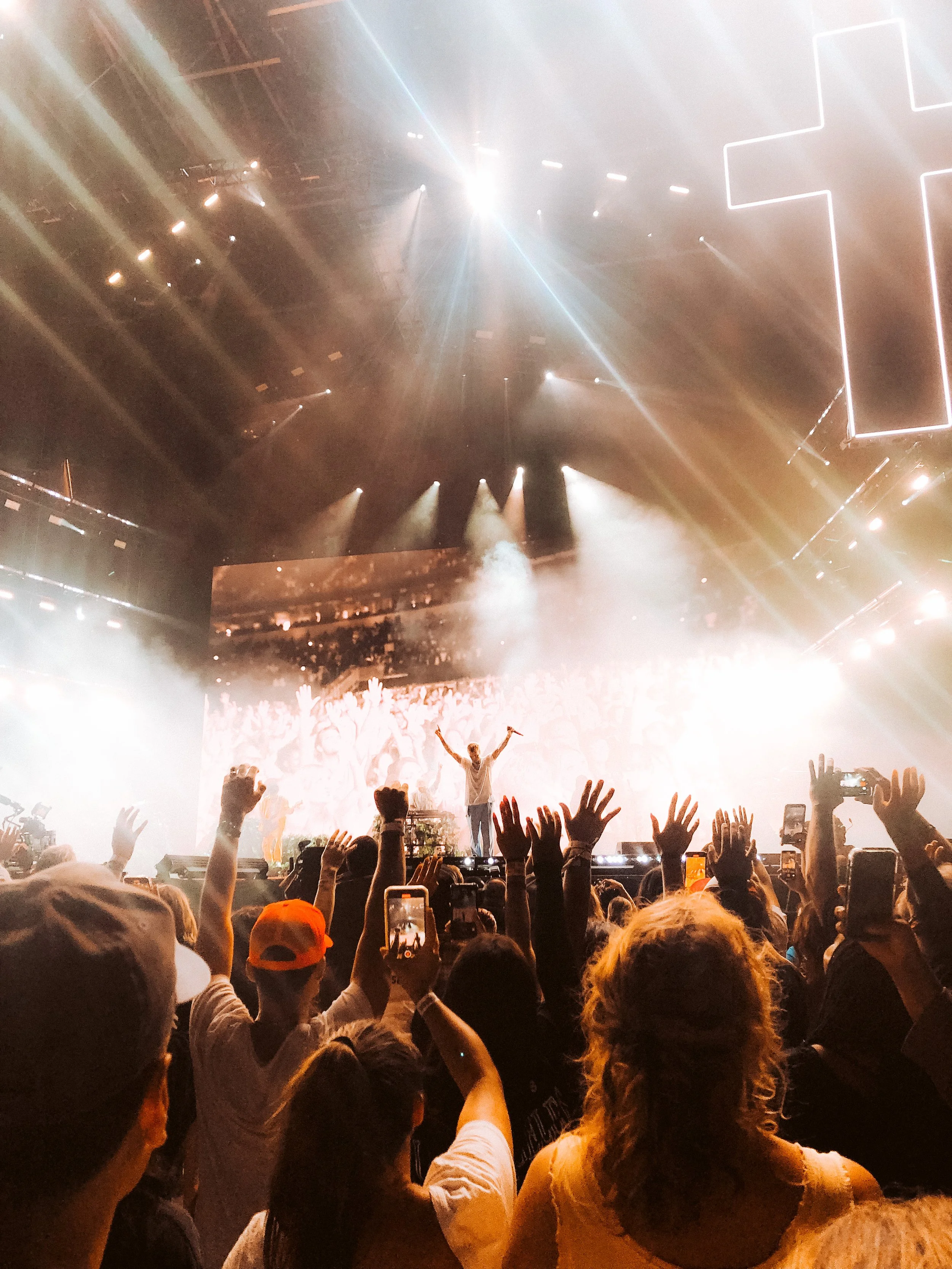 A concert with a performer on stage raising his arms, surrounded by bright lights and a large audience with many people raising their hands and taking photos.