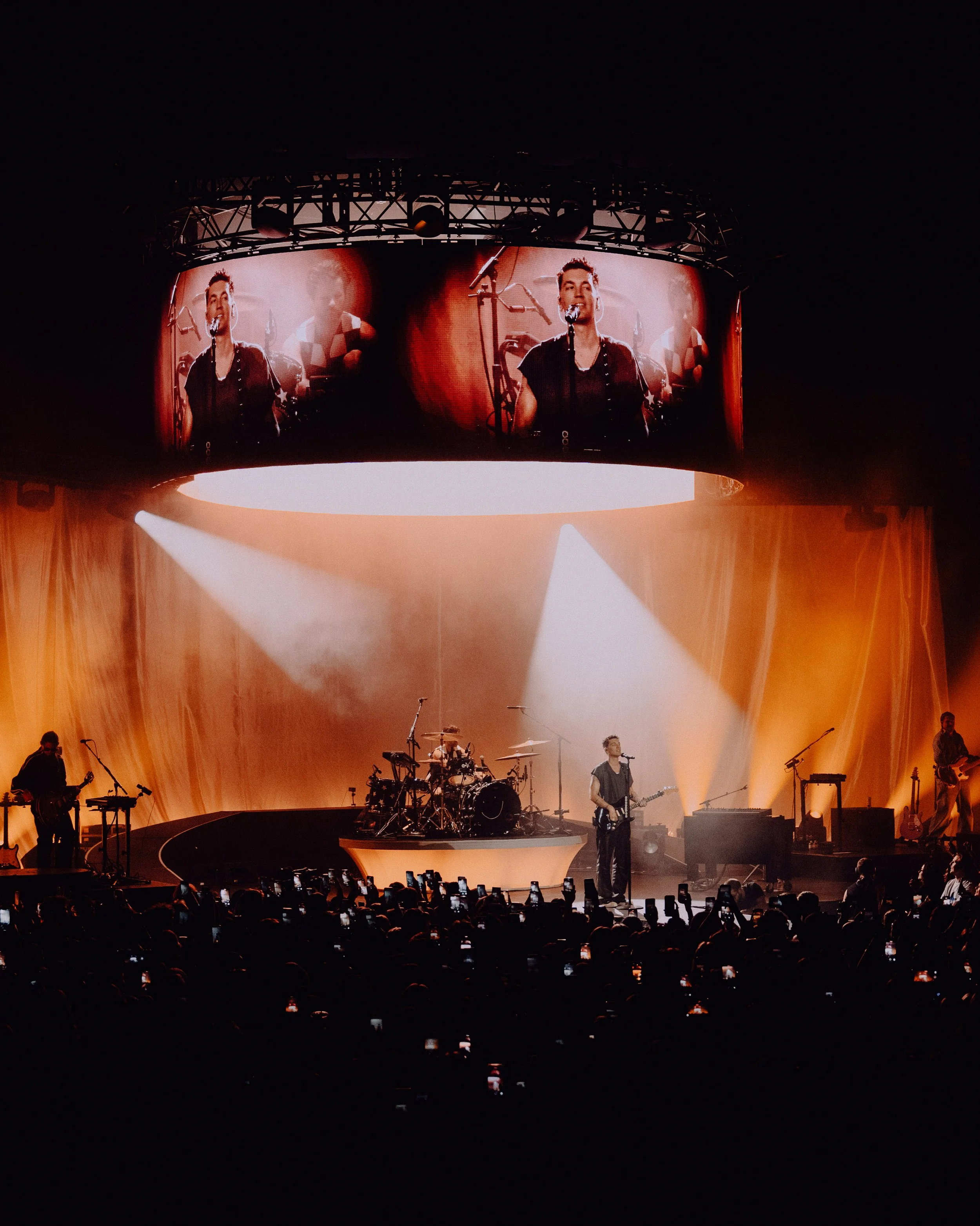 Concert stage with band performing under orange lighting, large screen overhead shows close-up of lead singer, audience taking photos.