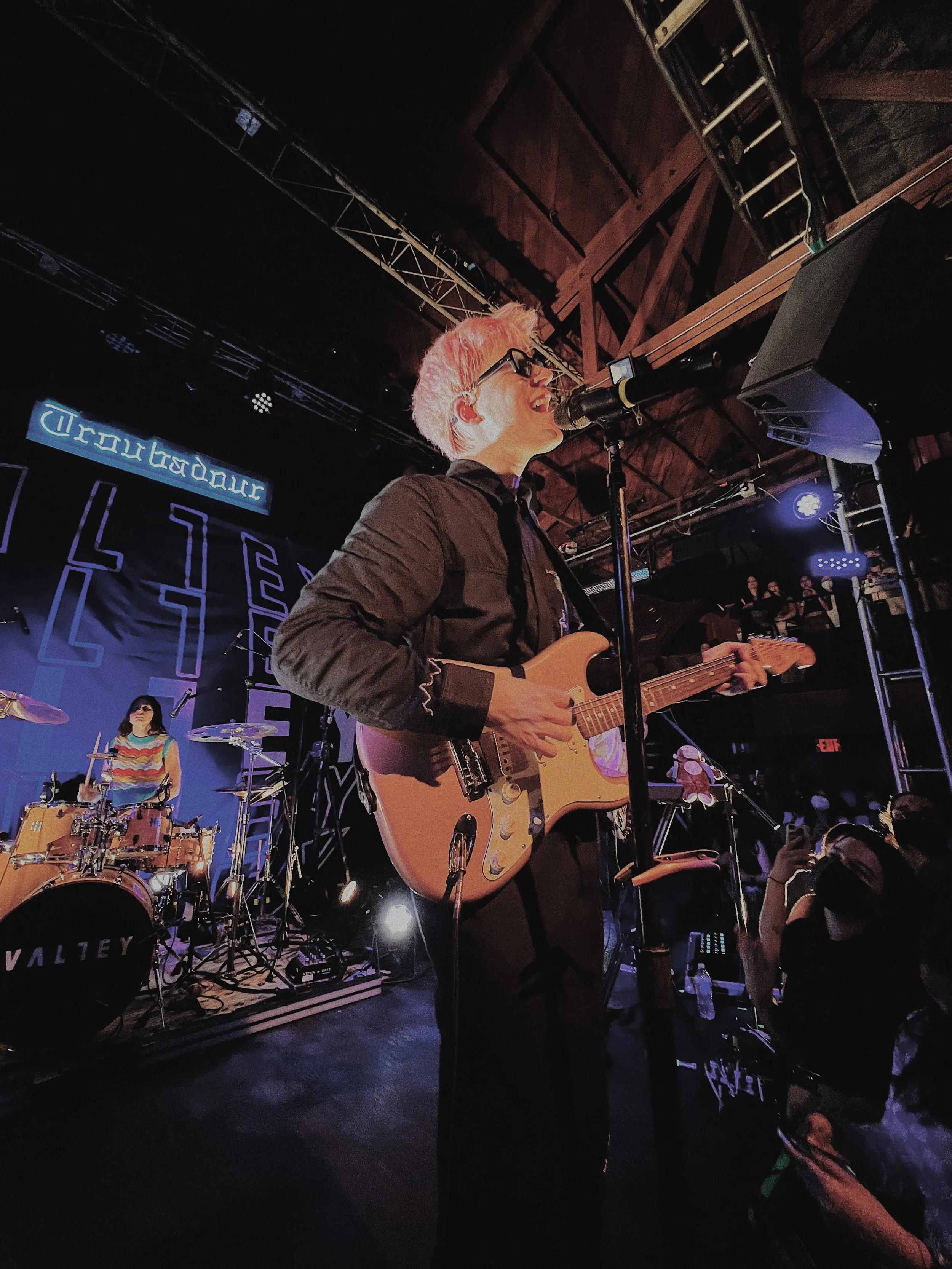 Musician with pink hair, glasses, and dark clothing playing an electric guitar and singing into a microphone on stage, with a drummer and a band logo in the background.