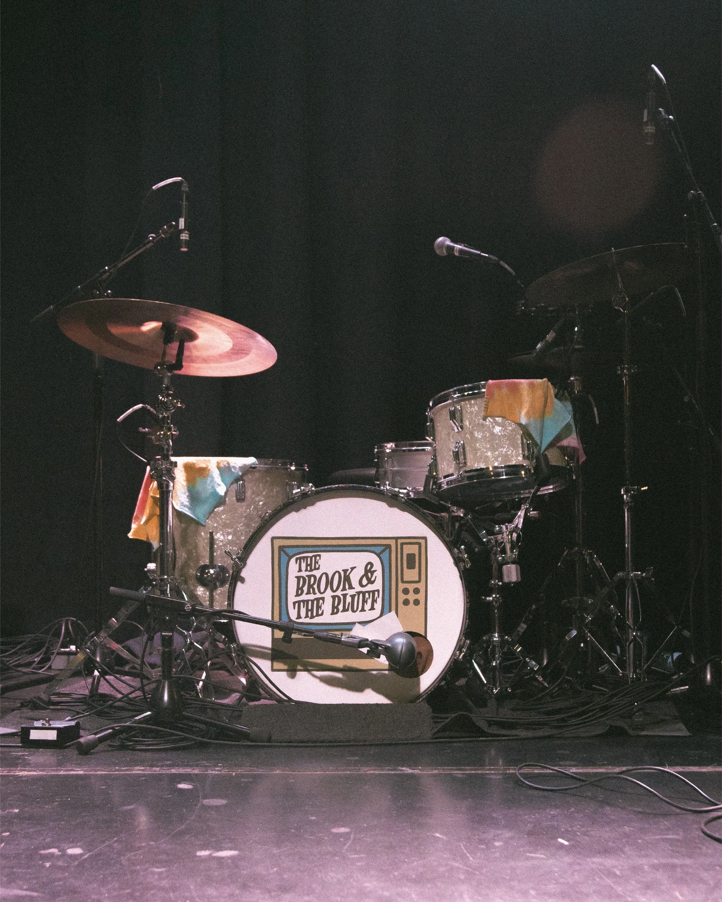 A drum set with a banner on the bass drum reading 'The Brook & The Bluff,' surrounded by microphones and cables, set on a stage with a dark background.
