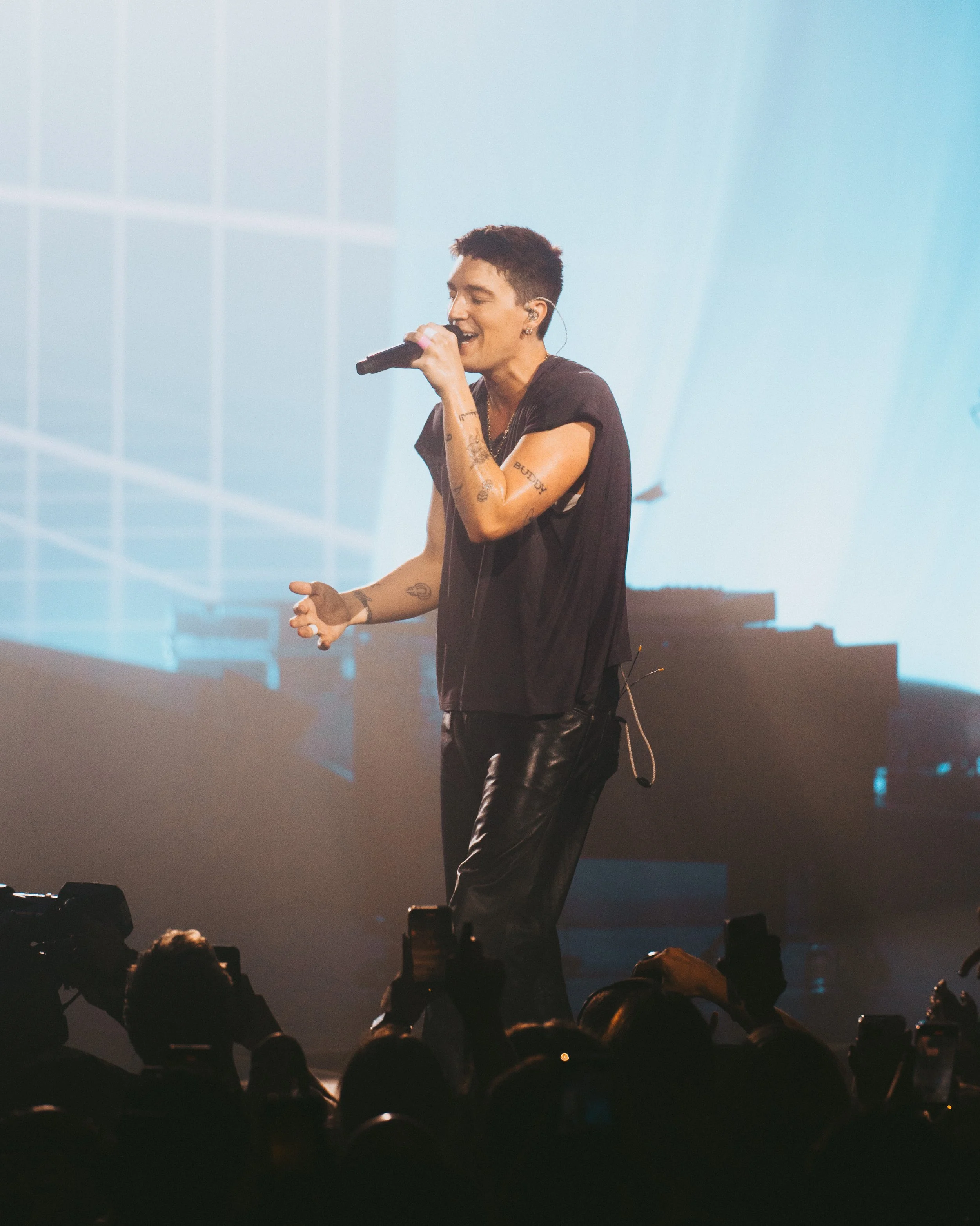 A singer performing on stage with a microphone, wearing a black t-shirt and leather pants, with tattoos on his arms, and an audience taking photos.