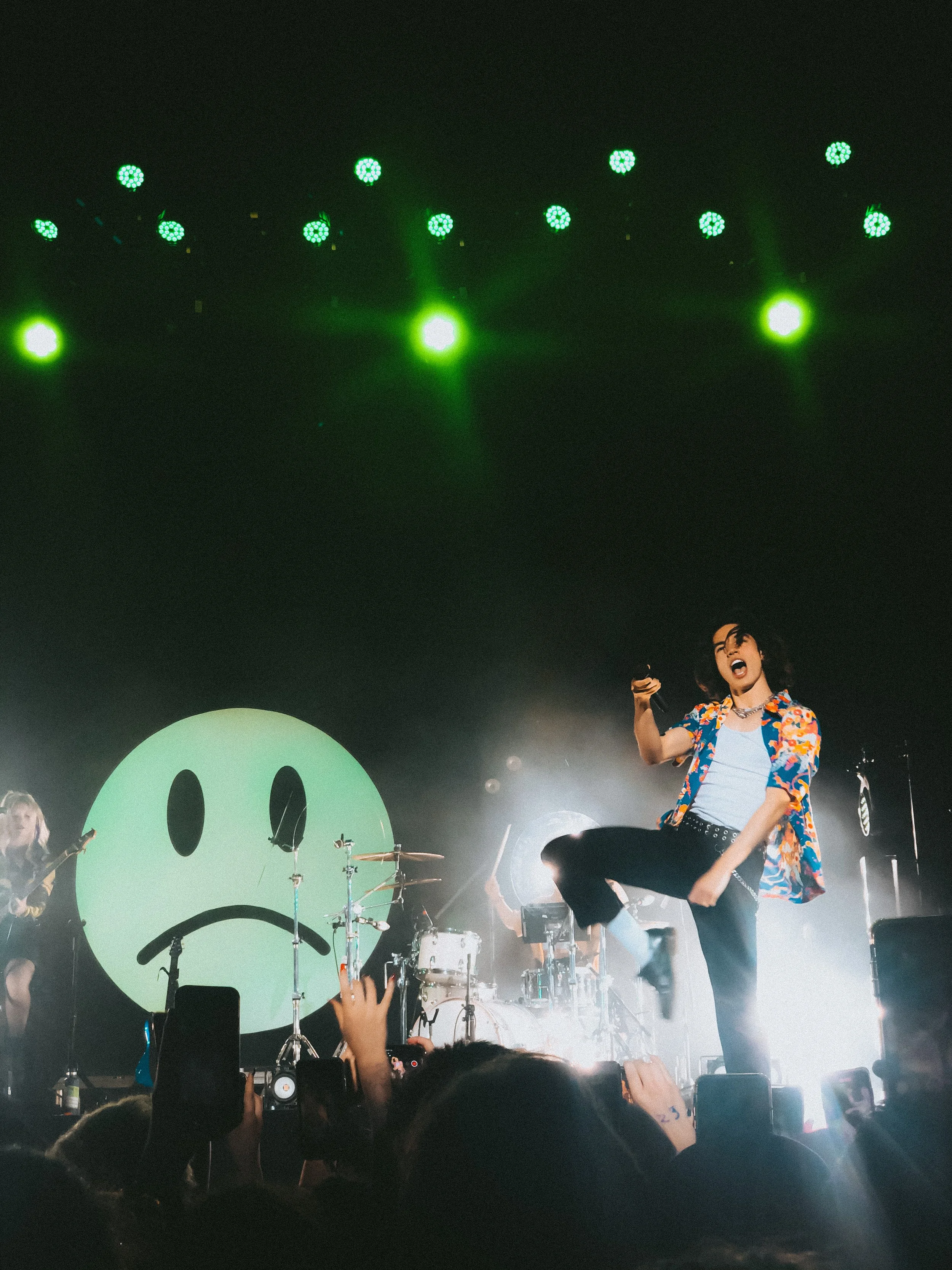 A live concert scene with a singer in a colorful shirt jumping in front of a large green smiley face on a screen, surrounded by band members and audience holding up phones.