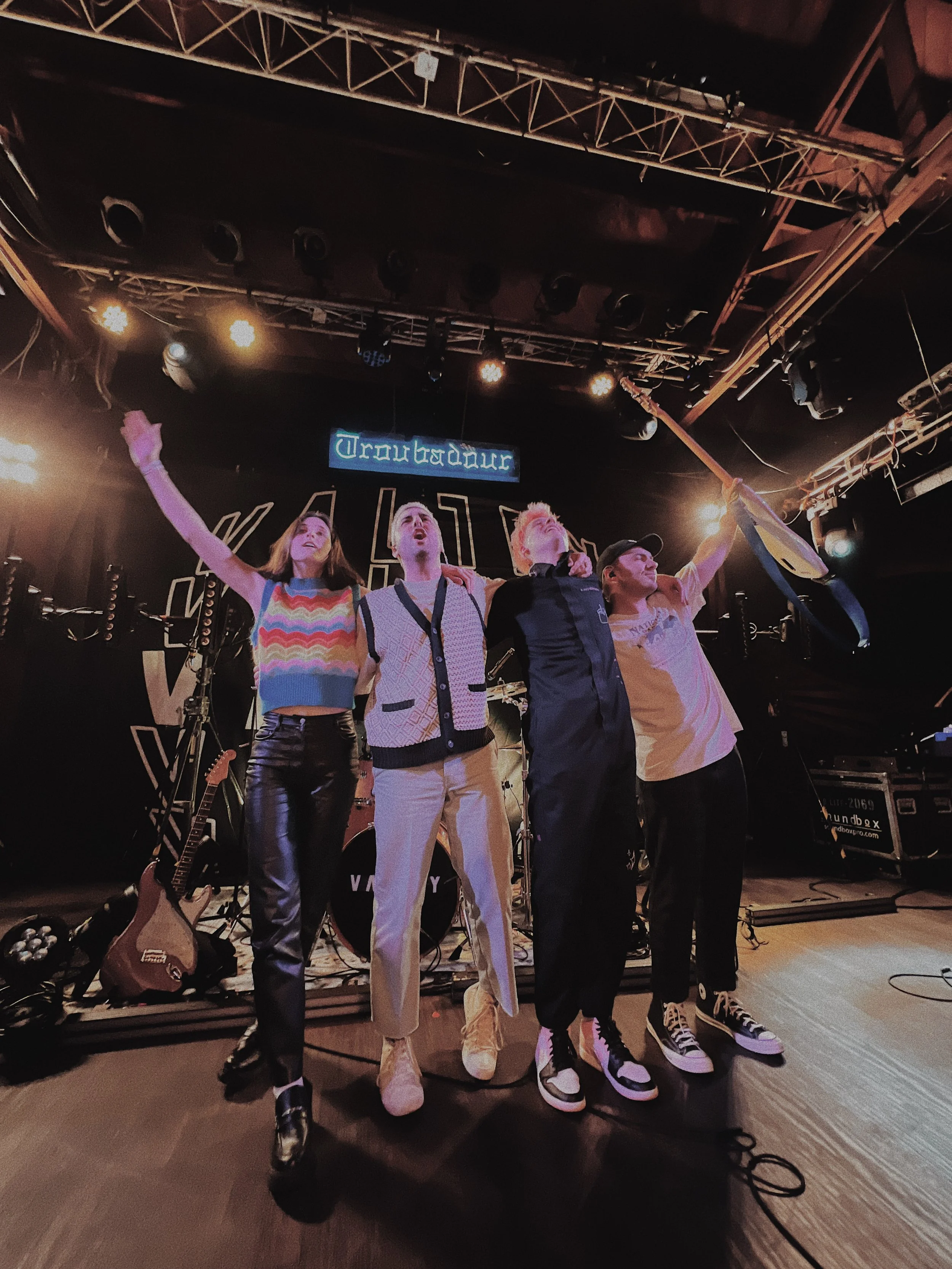 Four young bands stand on stage with their arms around each other after a performance, under stage lighting with a 'Troubadour' sign overhead, and musical instruments including a guitar and drum set behind them.