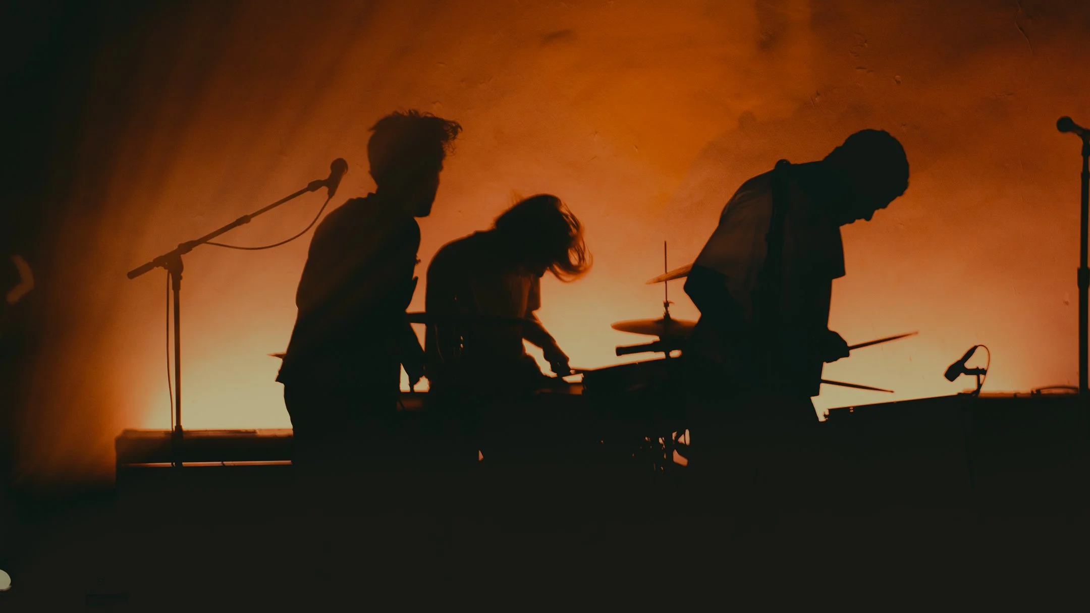Silhouettes of three musicians playing instruments on stage with warm orange lighting in the background.