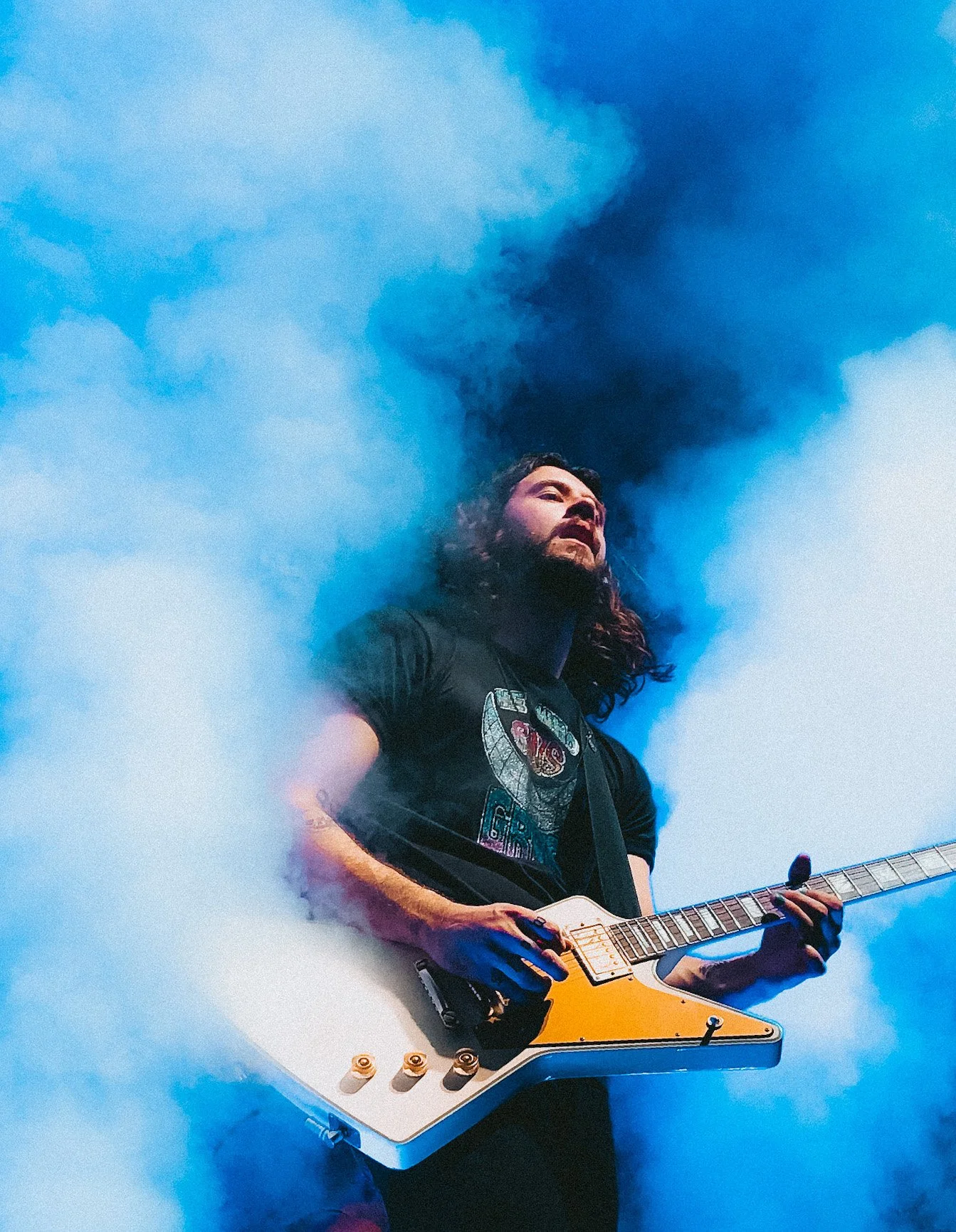 A male guitarist with long, curly hair and a beard plays an electric guitar amidst blue smoke on stage.