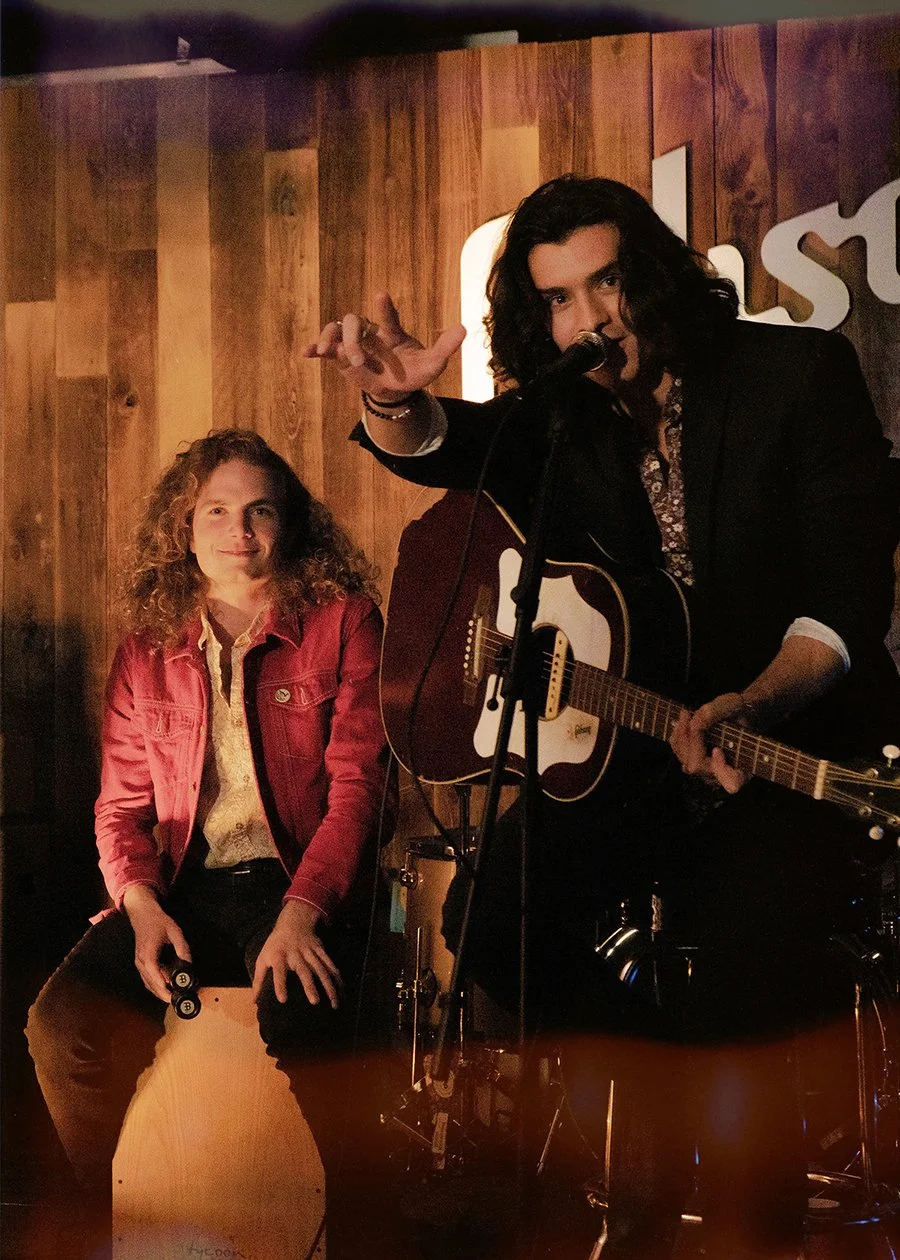 A young woman with curly hair in a red jacket sitting next to a man with long dark hair singing and playing an acoustic guitar at a music venue with wooden walls.