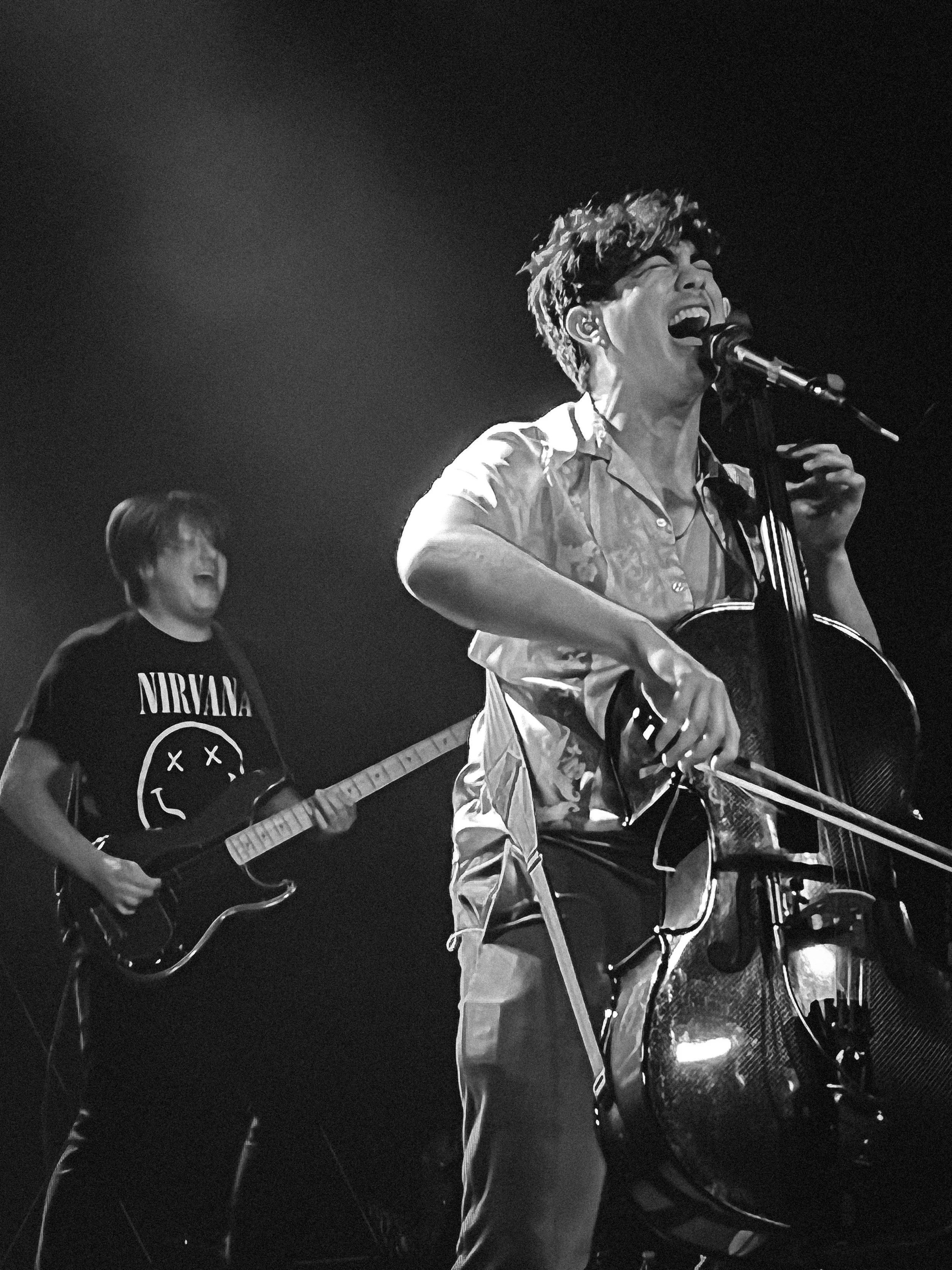 Two musicians performing on stage: one singing passionately into a microphone and playing a cello, and the other in the background playing an electric guitar and wearing a Nirvana T-shirt, both enjoying the performance.