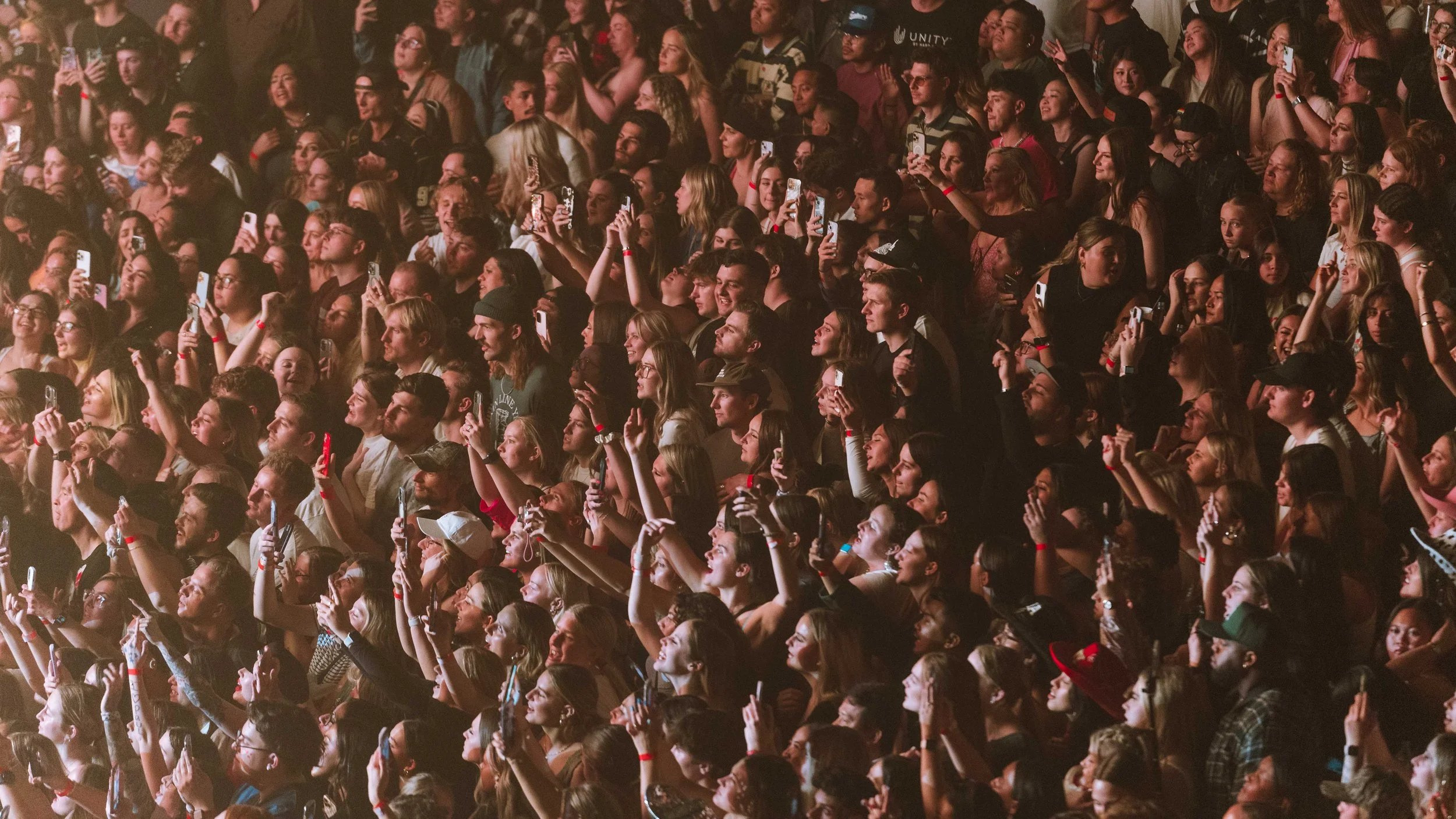 Large crowd of people at a concert or event, many holding up phones taking pictures, with some smiling, cheering, and enjoying the moment.
