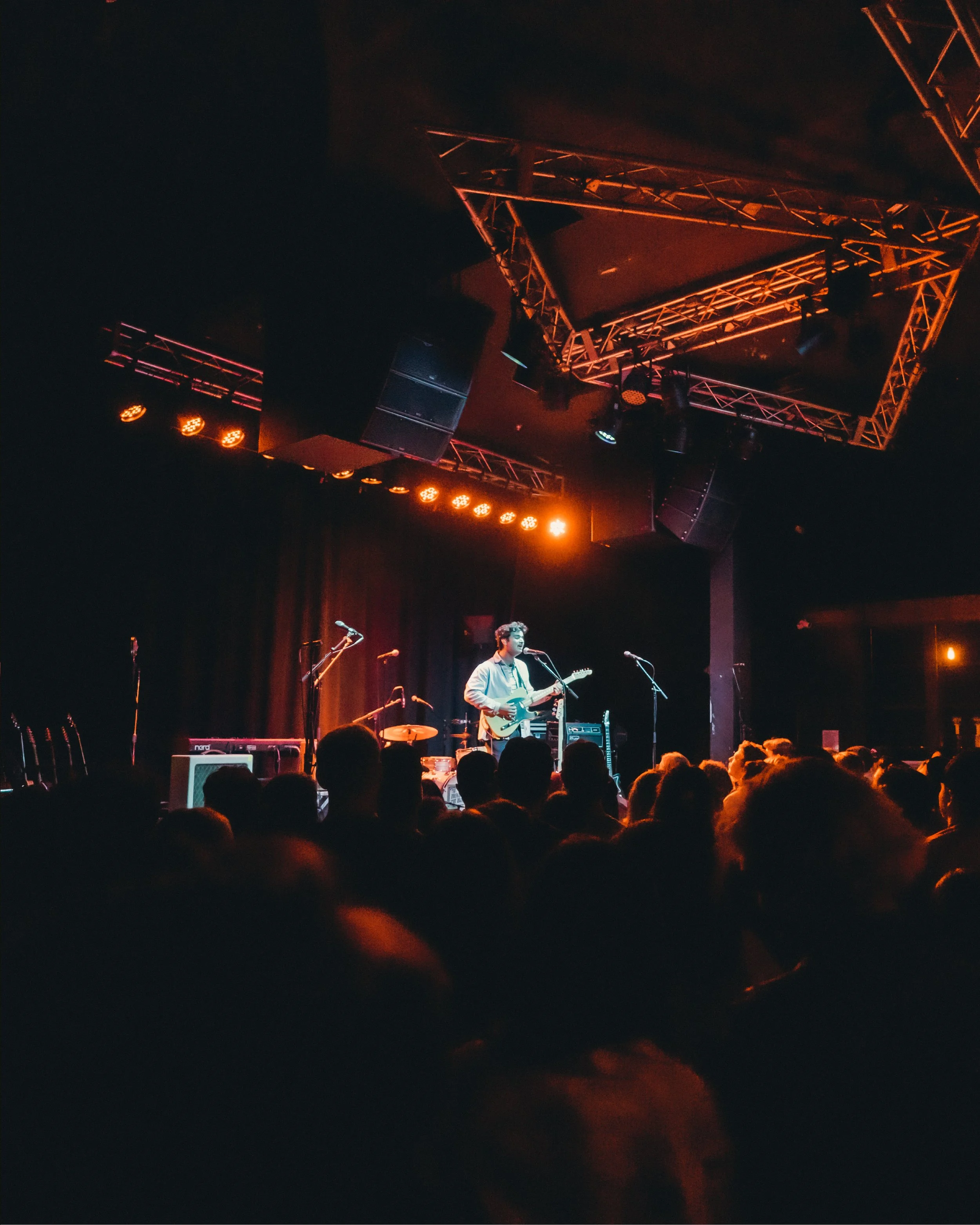 A musician performing on stage with guitar, surrounded by stage lights and audience in a dark concert venue.