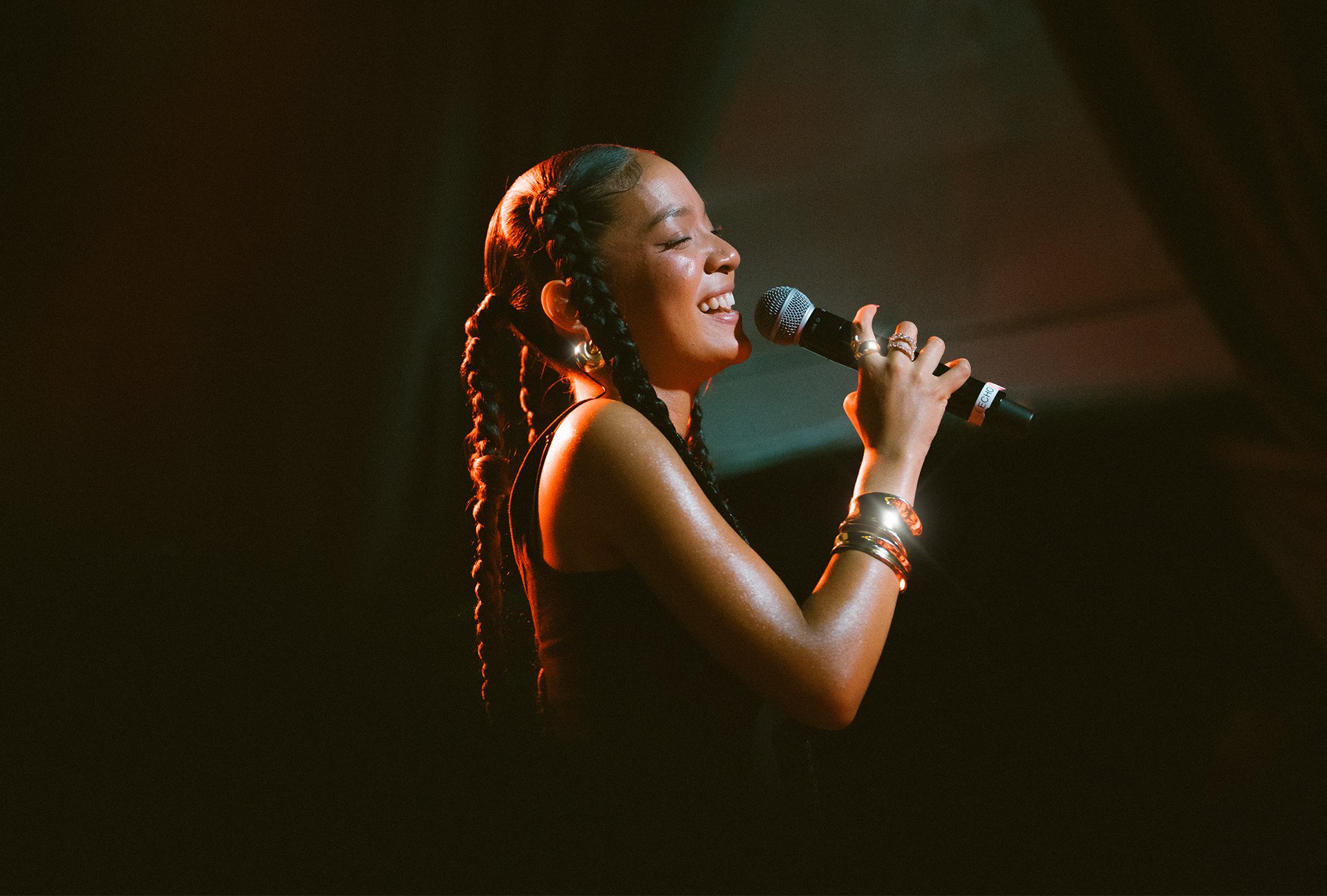 A woman singing into a microphone on stage, with braided hair, wearing a sleeveless top and multiple bracelets, smiling with her eyes closed.