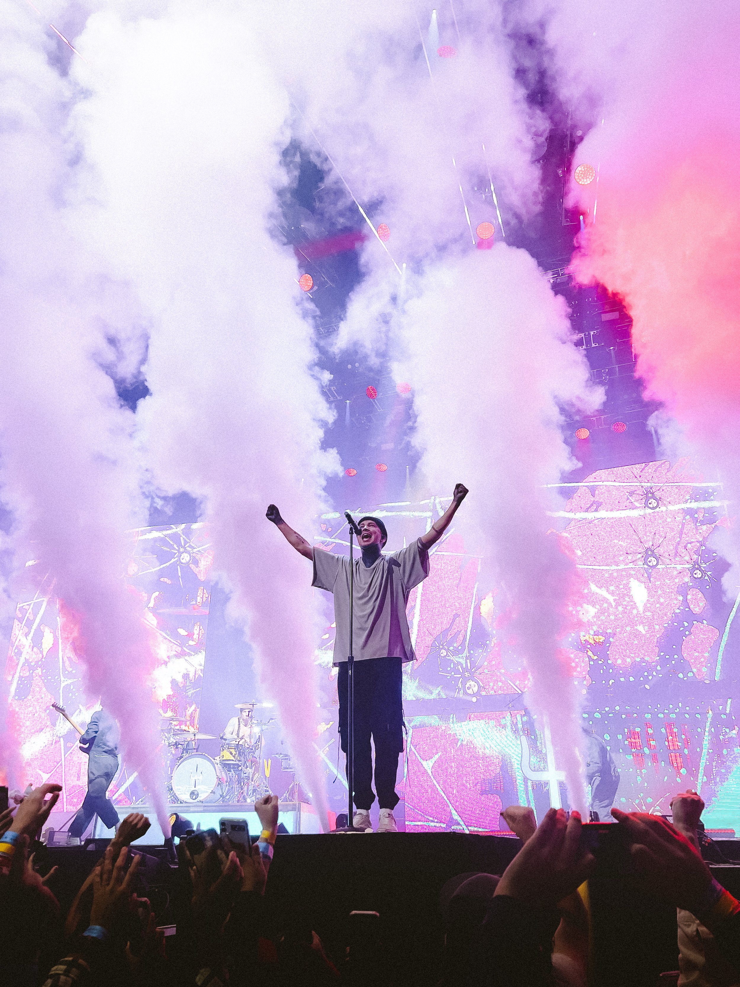 A performer on stage with arms raised, surrounded by colorful smoke, with a live band playing behind them and an audience in front.