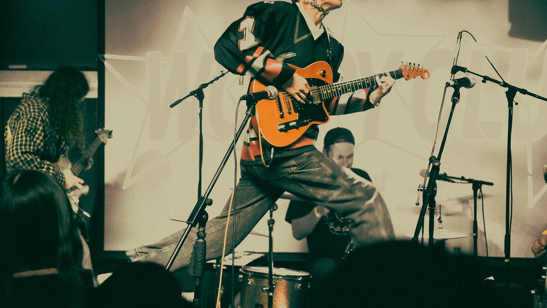 Musicians performing on stage, with one playing an electric guitar while jumping, others playing instruments in the background, microphones and music stands set up, audience visible at the bottom.