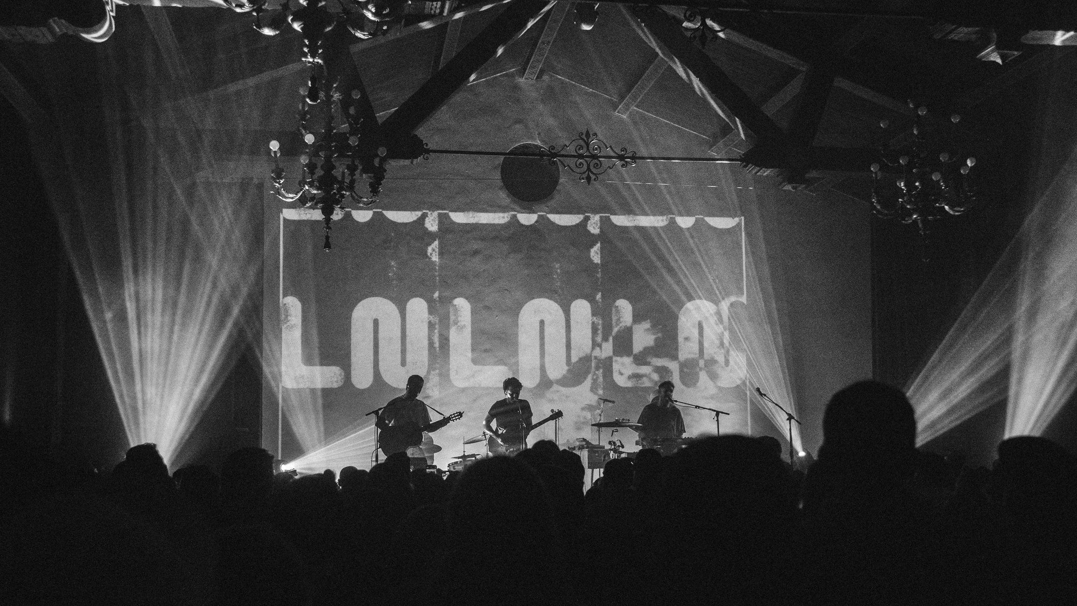 Black and white photo of a live band performing on stage, with three musicians playing guitars and drums, in front of a large screen displaying the word 'LULLABO.' The audience is visible in the foreground, and ornate chandeliers hang from the ceiling.