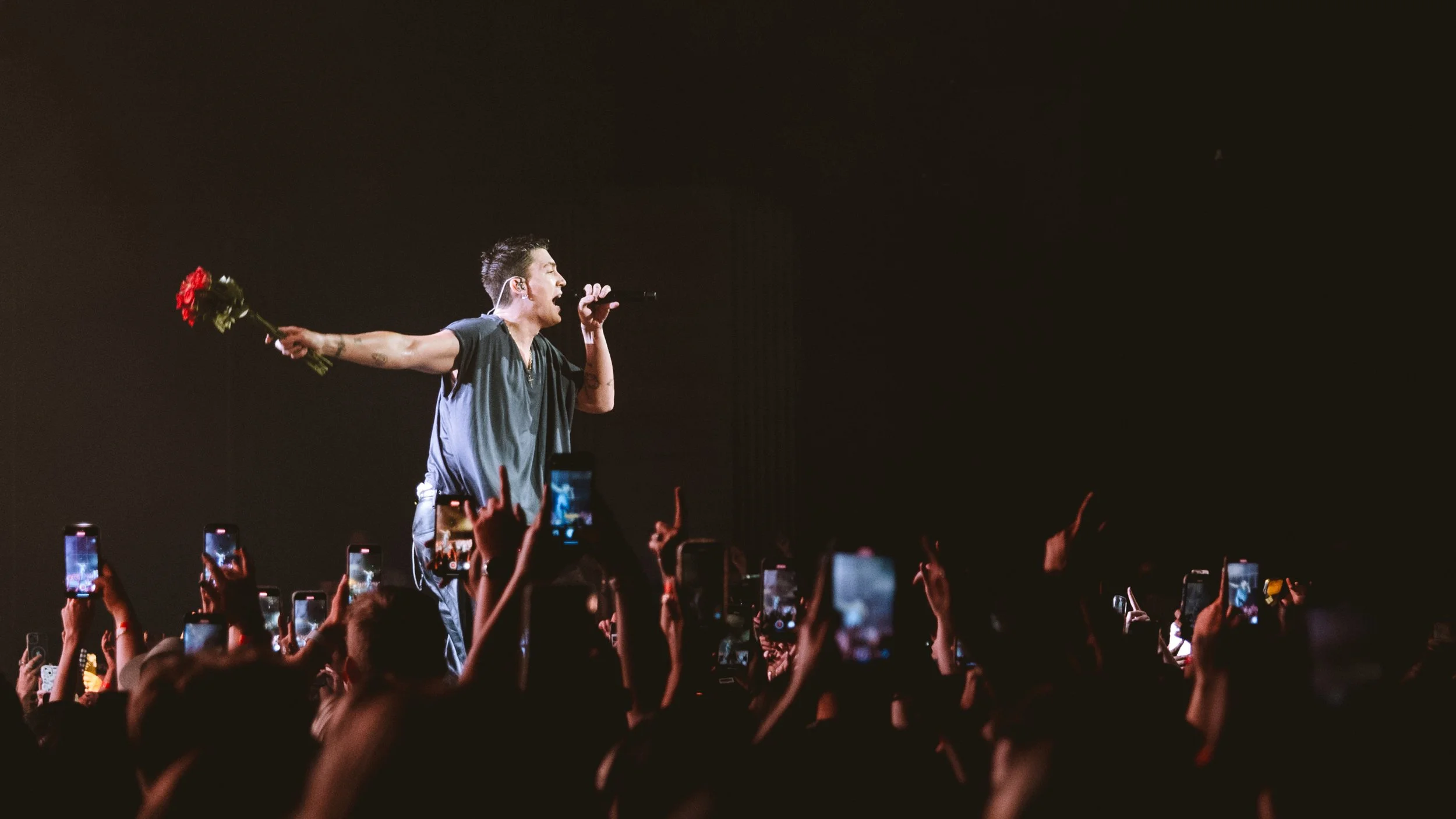 Performer on stage holding a microphone in one hand and a bouquet of red roses in the other, with a dark background and audience members taking photos with their phones.