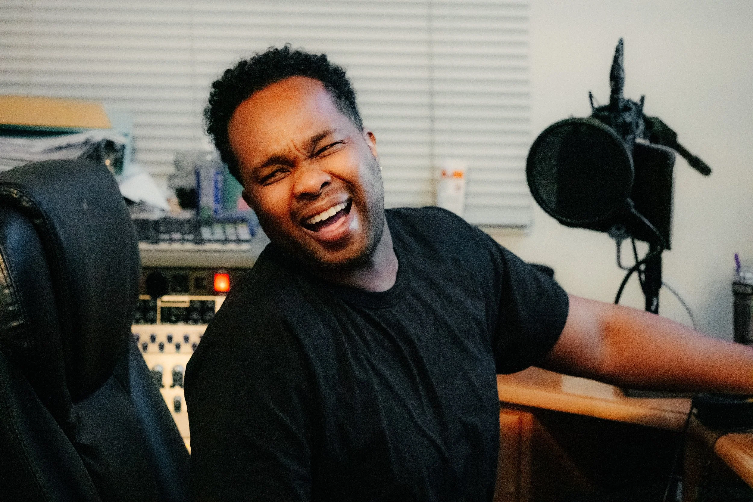 A man with black curly hair and a beard laughing with mouth open in a recording studio or music production environment.