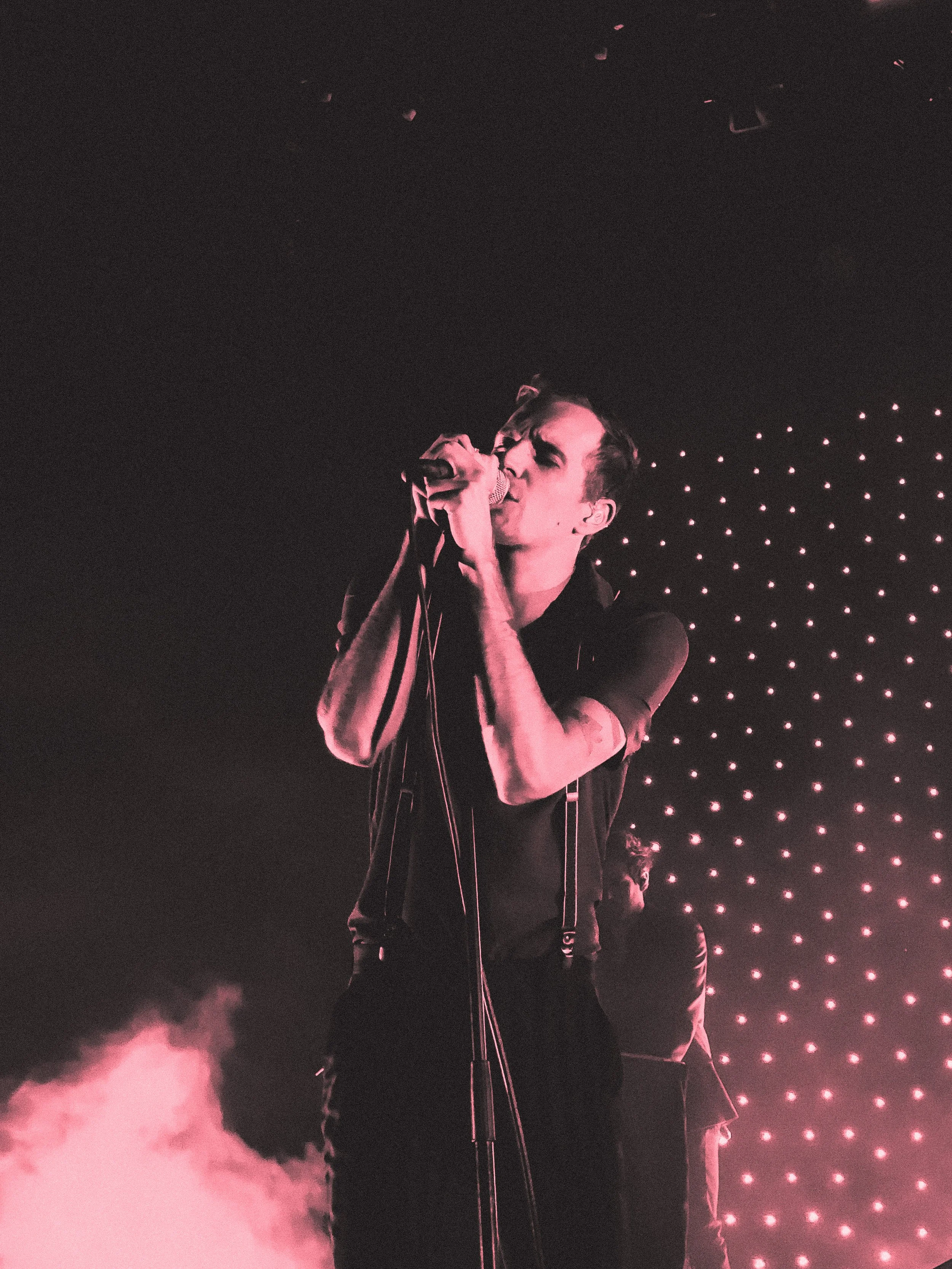 A male singer performing on stage with a microphone, pink stage lighting, and a dotted LED background.