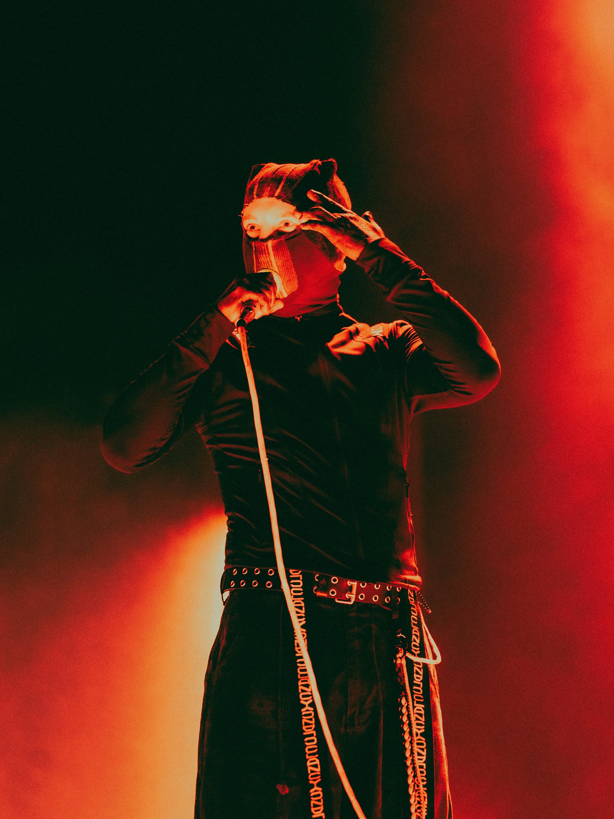 A person wearing a mask, a hooded jacket, and a studded belt, holding a microphone on a stage with red and orange lighting.