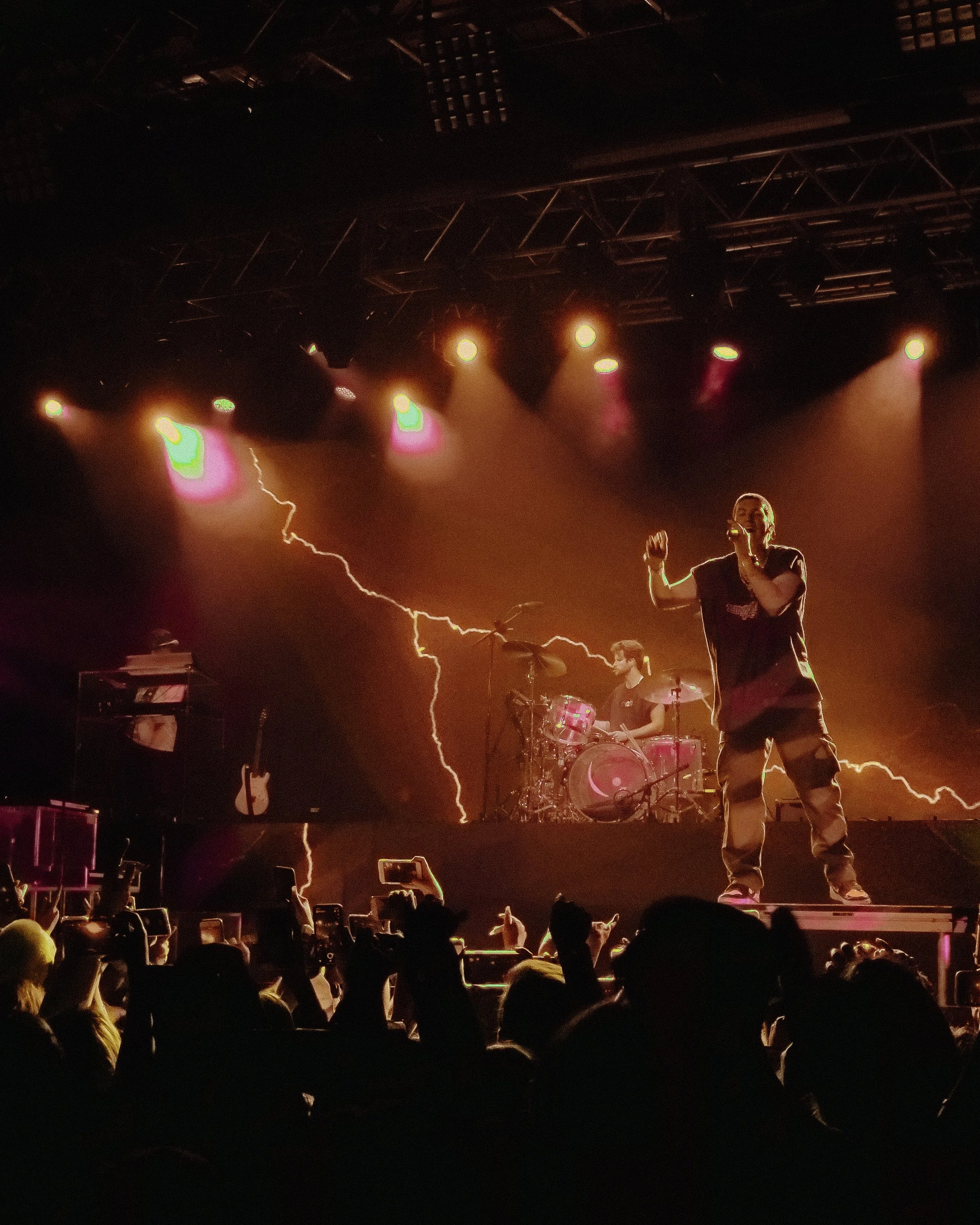 A live concert performance with a singer on stage, a drummer in the background, colorful stage lighting, and a lightning effect on a large screen behind them, with an audience watching and recording.