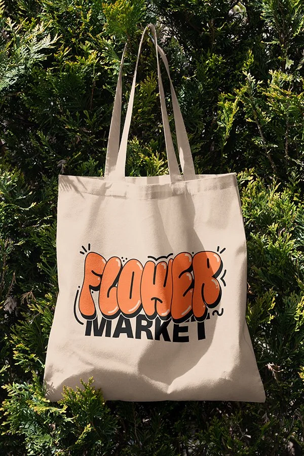 A beige tote bag with the words "Foucher Market" printed on it, hanging from a plant bush.