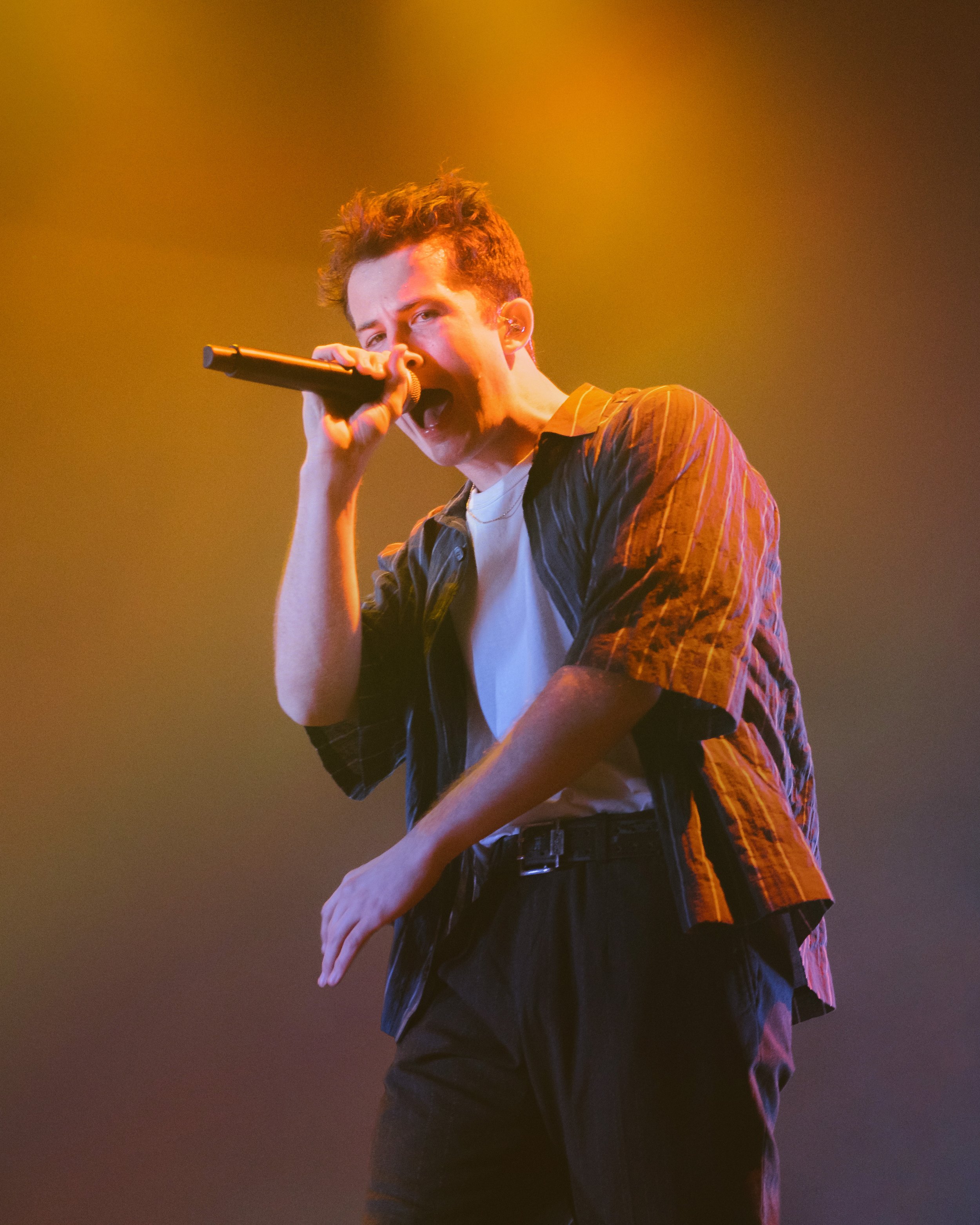 Young man singing passionately into a microphone on stage, wearing a striped shirt and white t-shirt, with colorful stage lighting.