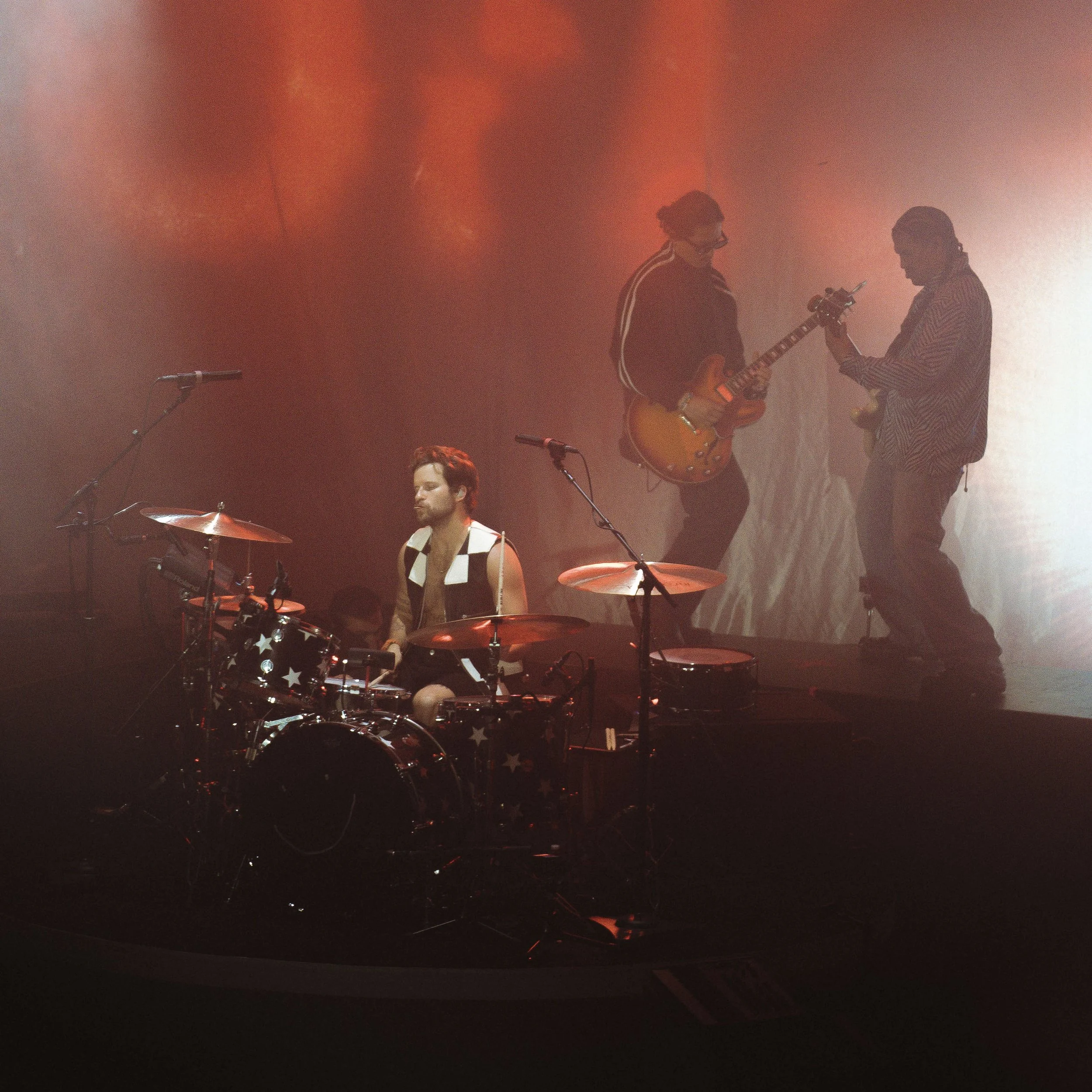 Three musicians performing on stage. One is seated at drums with star patterns, and two are standing, playing electric guitars, with a reddish stage lighting background.